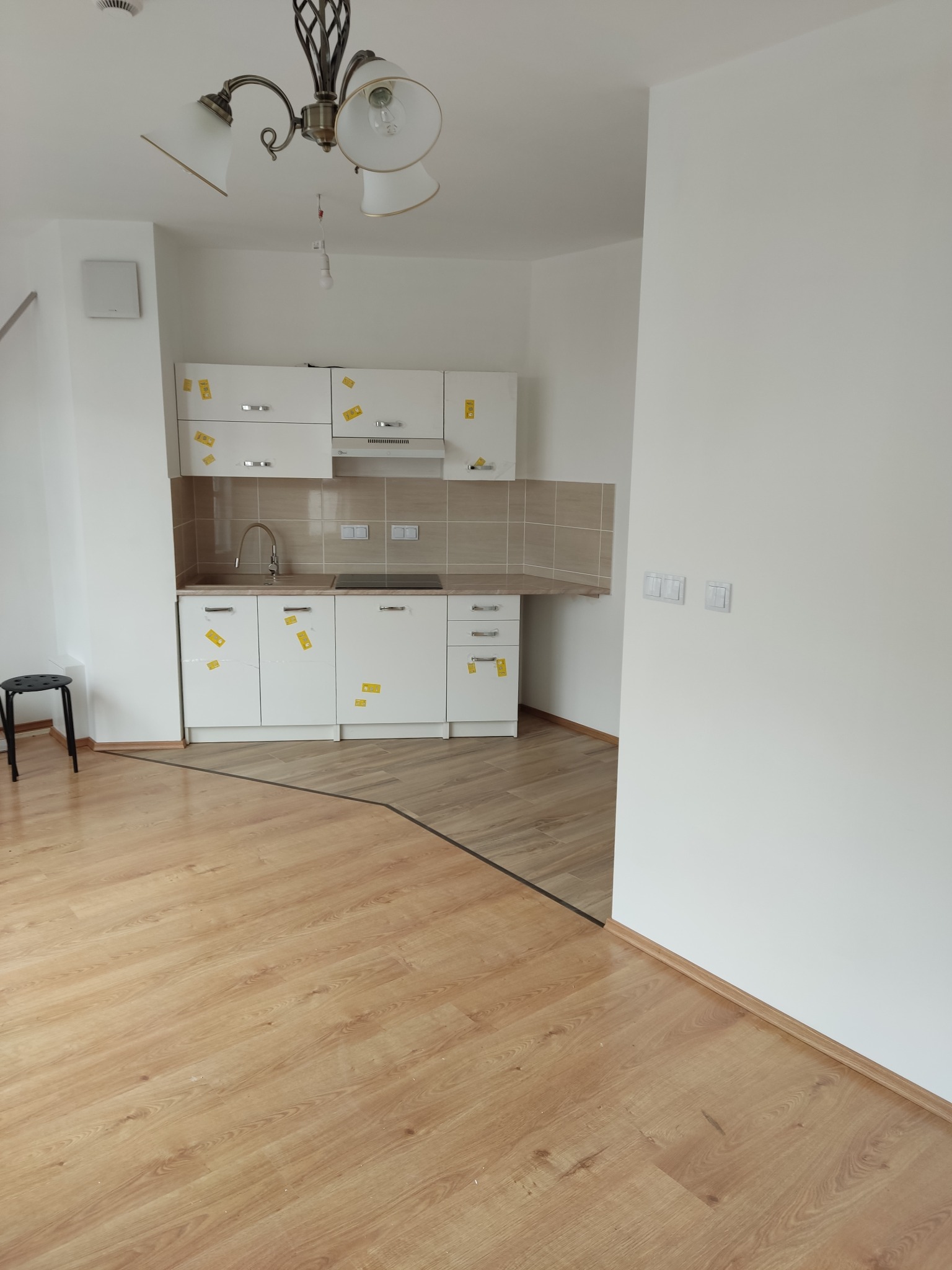 Jasna kuchnia z drewnianą podłogą w Konstancin-Jeziorna. Białe szafki z naklejkami zabezpieczającymi. Widoczny fragment podłogi i krzesło.