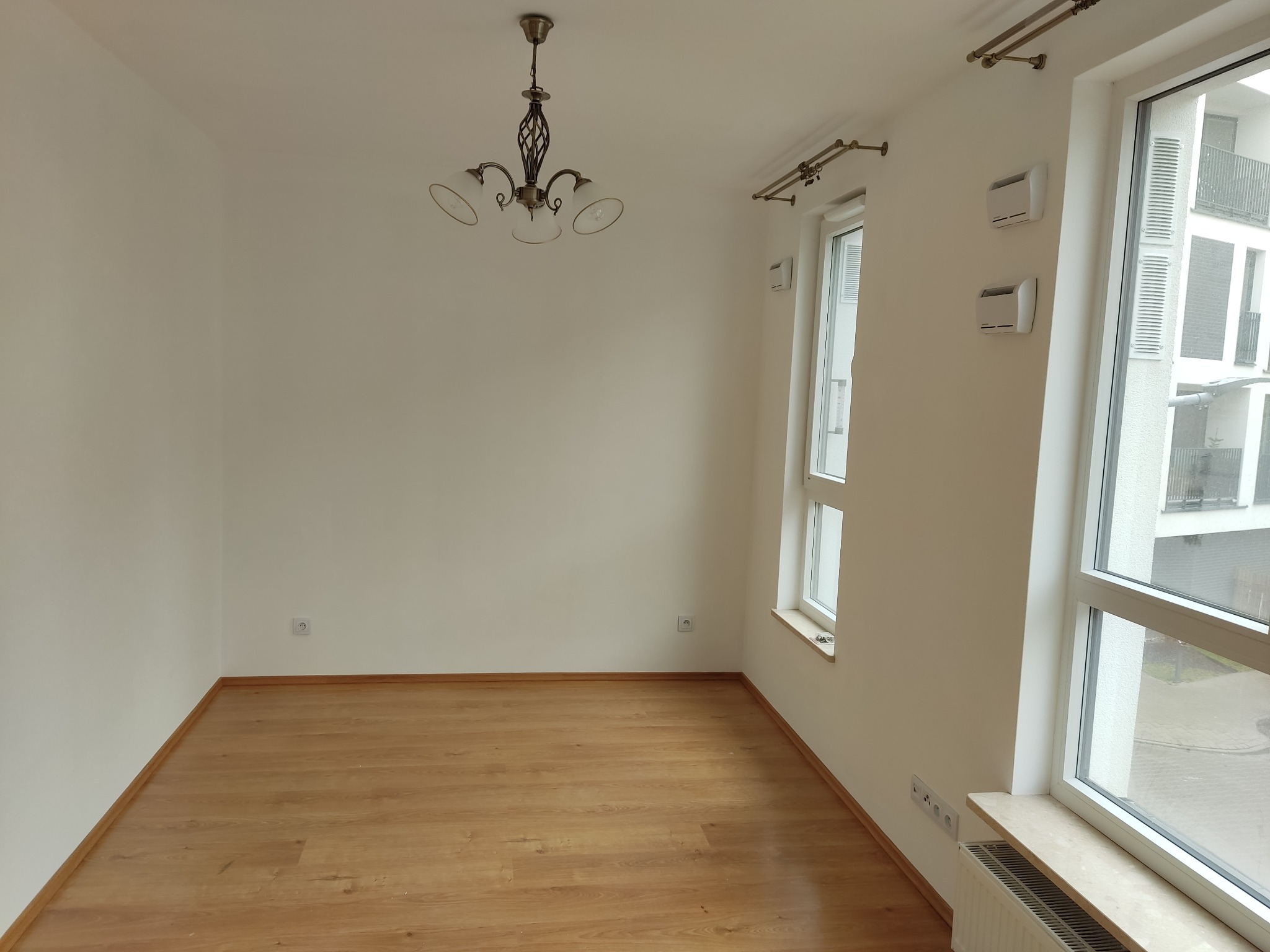 Jasne, puste wnętrze z drewnianą podłogą, białymi ścianami i oknami z widokiem na balkon. W centralnej części sufitu ozdobny, metalowy żyrandol.