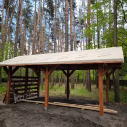 Wznoszona drewniana konstrukcja z beżowym pokryciem dachowym na tle leśnym. Widoczne elementy konstrukcyjne i drabina. Częściowo zabudowana ściana z desek.