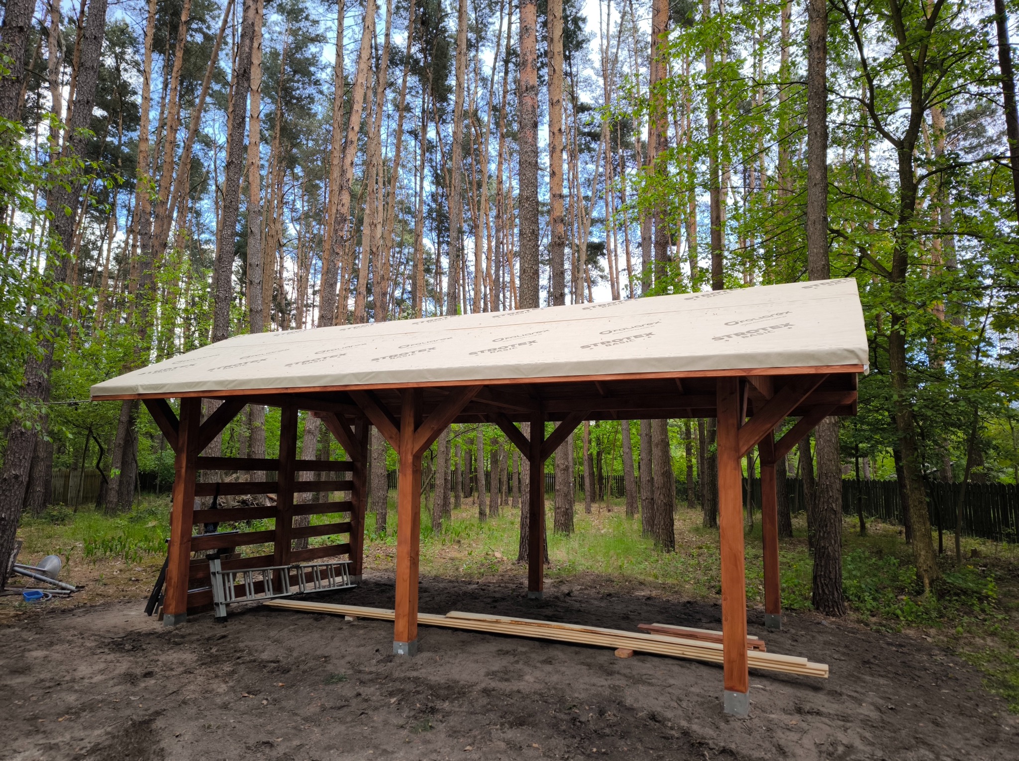 Wznoszona drewniana konstrukcja z beżowym pokryciem dachowym na tle leśnym. Widoczne elementy konstrukcyjne i drabina. Częściowo zabudowana ściana z desek.