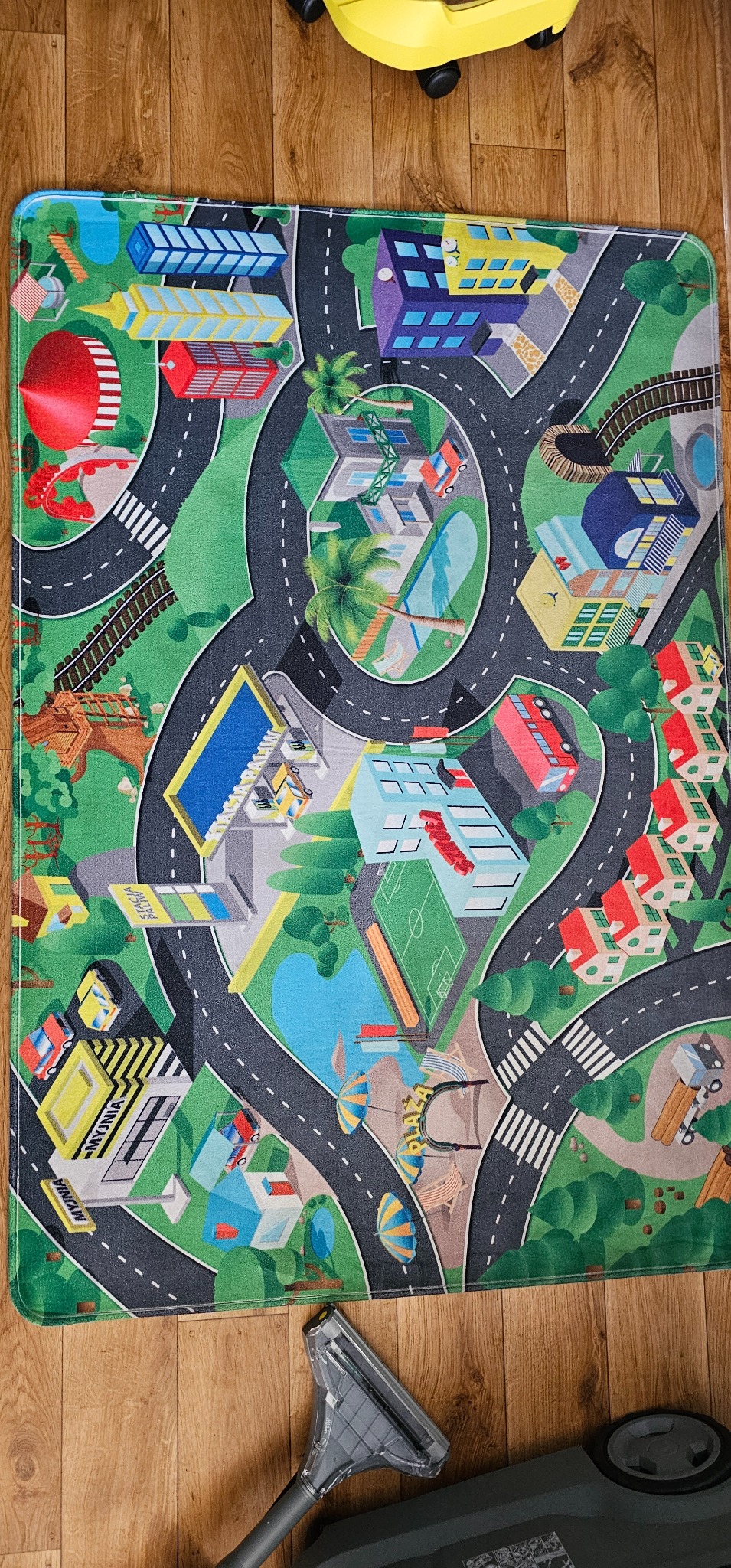 Kolorowy dywan z motywem miasta, leżący na drewnianej podłodze, częściowo przysłonięty przez końcówkę odkurzacza i fragment żółtego urządzenia czyszczącego.