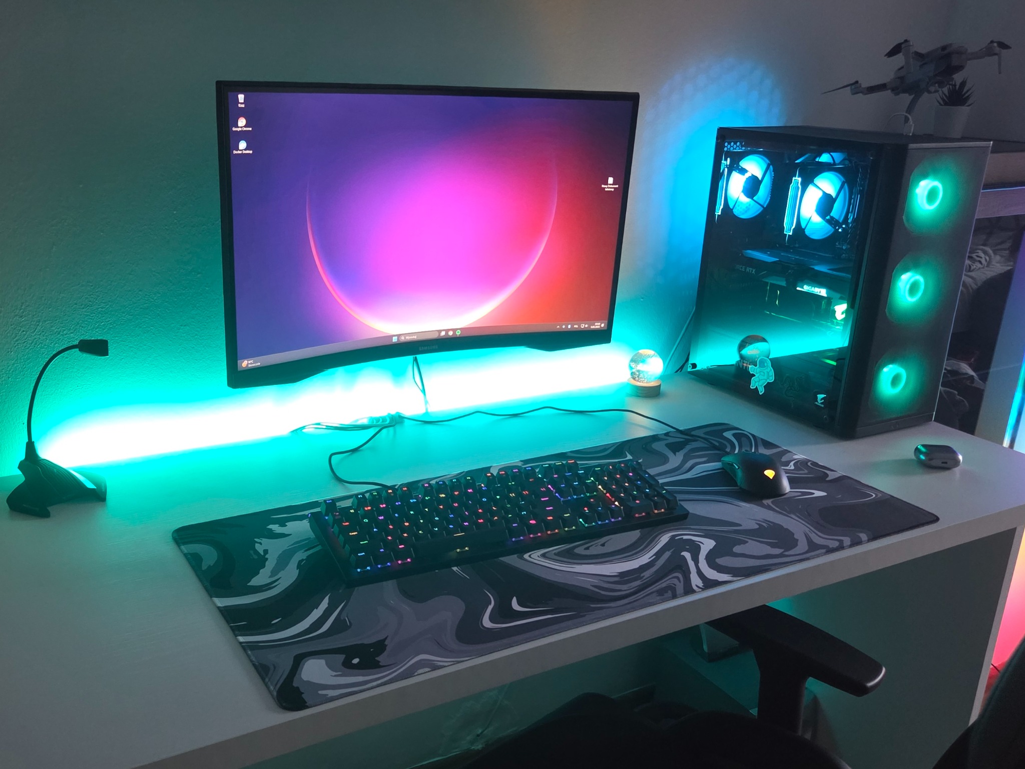 Komputer stacjonarny z podświetleniem LED, monitor na biurku z podkładką pod mysz. Widoczna klawiatura i mysz. Na obudowie komputera stoi model drona.