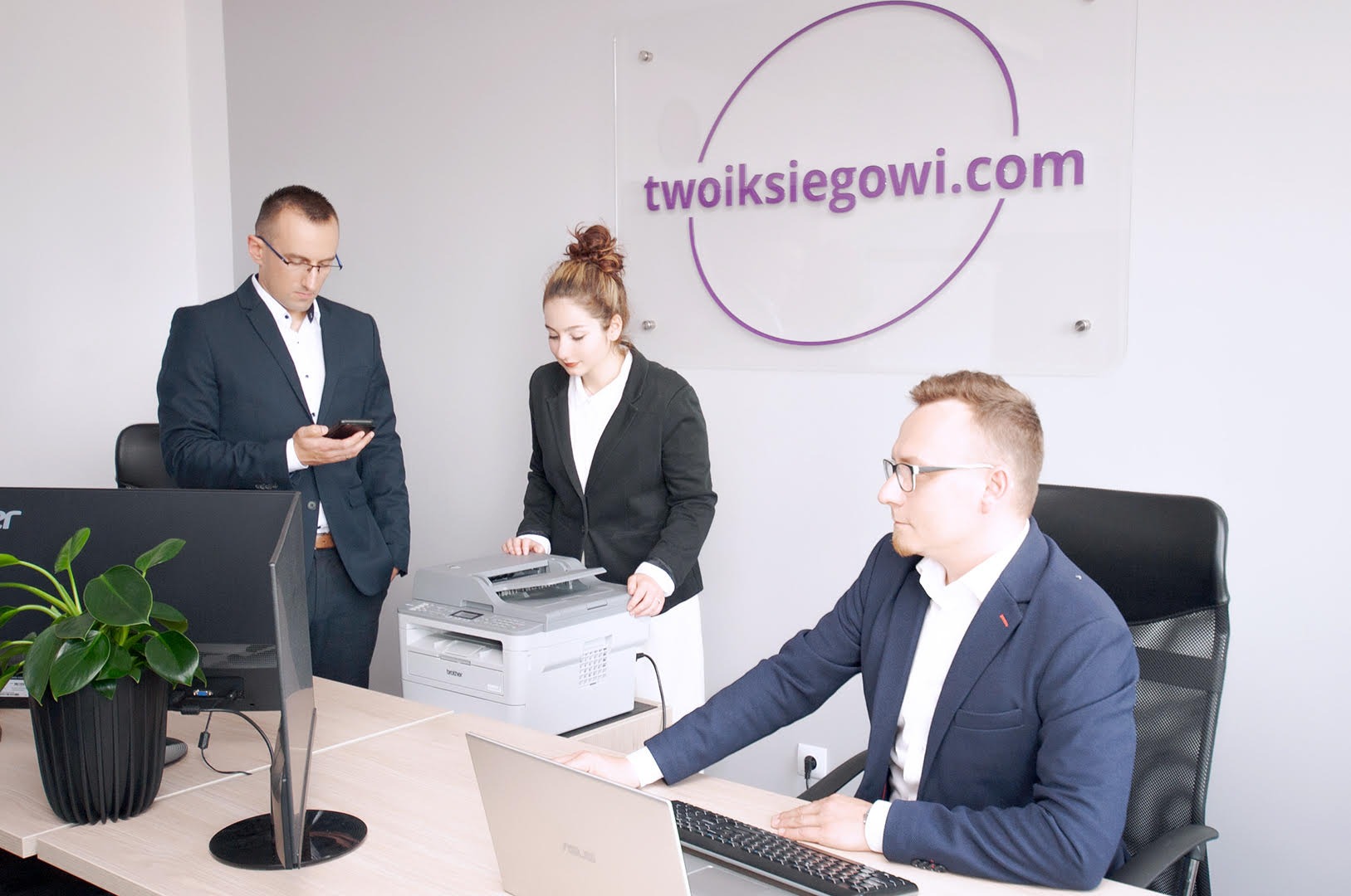 Biuro księgowe: trzech pracowników w garniturach przy biurku z laptopem i drukarce, logo firmy widoczne na szklanej tablicy na ścianie.