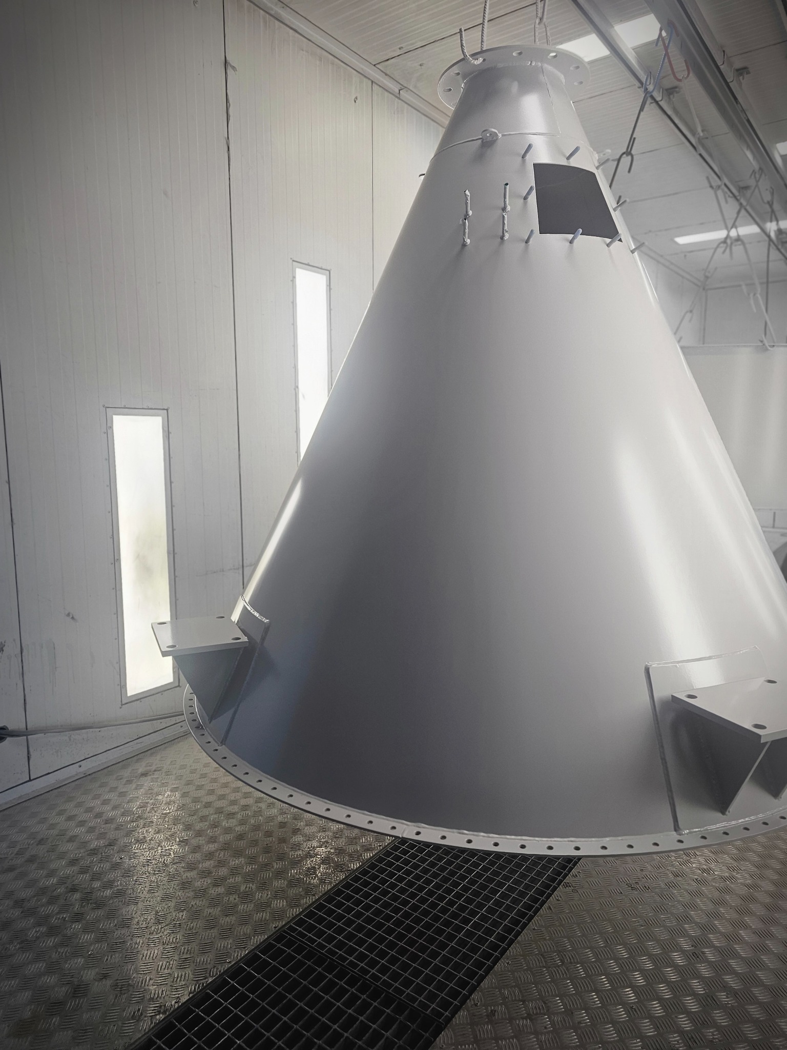 Stożkowy, metalowy element w jasnej kabinie lakierniczej. Widoczne otwory i uchwyty montażowe. Podłoga z kratką i blachą ryflowaną. Oświetlenie z boku.