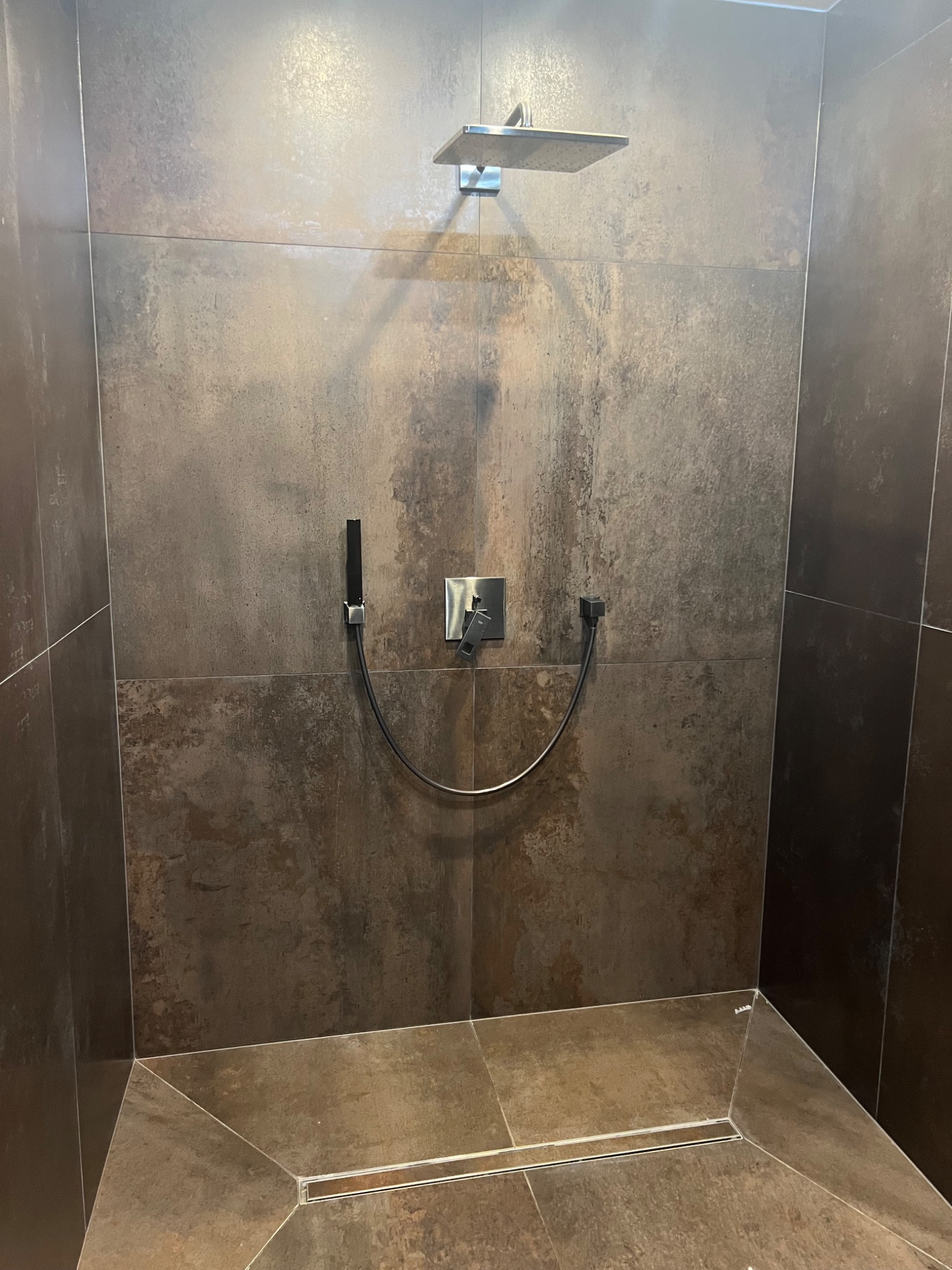 Nowoczesna kabina prysznicowa z deszczownicą i odpływem liniowym, wykończona płytkami w odcieniach brązu i szarości, tworząc elegancki i minimalistyczny design.