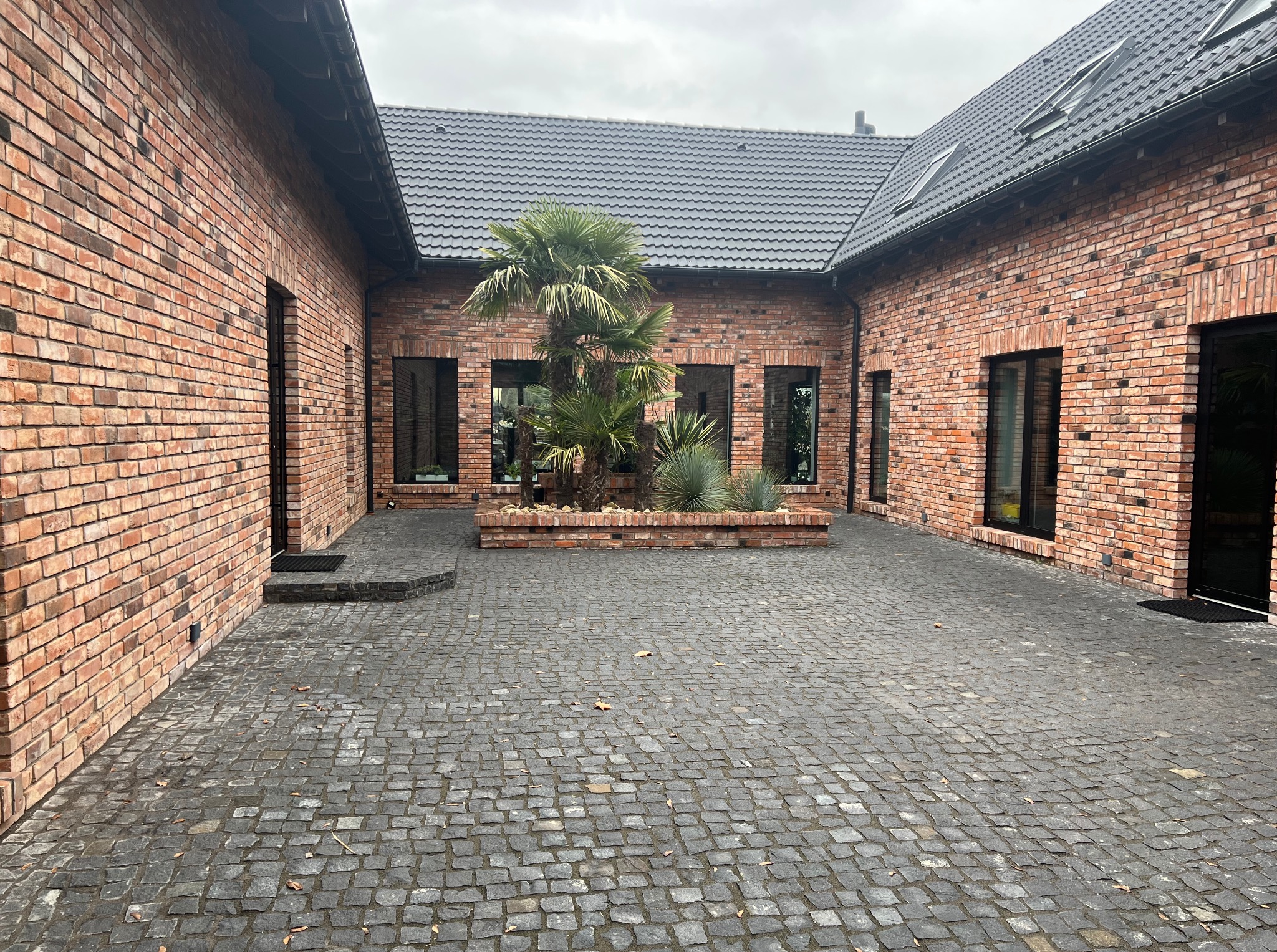 Dziedziniec domu z cegły klinkierowej, wyłożony kostką brukową. Na środku rabata z palmami i roślinami. Widoczne okna z czarnymi ramami i dach z dachówki.