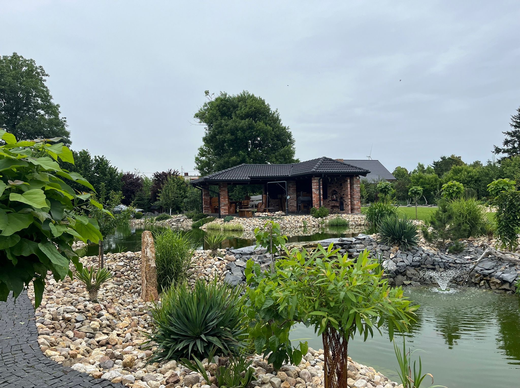 Ogród z oczkiem wodnym, kamieniami i murowaną altaną z ciemną dachówką w tle. Woda odbija niebo. Dekoracyjne rośliny na pierwszym planie.