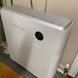 GREEN LEAF POWER SPÓŁKA Z OGRANICZONĄ ODPOWIEDZIALNOŚCIĄ - Biały, nowoczesny magazyn energii SiGenergy z okrągłym panelem sterowania, ustawiony w pomieszczeniu z widocznymi rurami i instalacjami na ścianie.