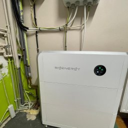 GREEN LEAF POWER SPÓŁKA Z OGRANICZONĄ ODPOWIEDZIALNOŚCIĄ - Biały magazyn energii Sienergy z podłączonymi kablami, umieszczony w pomieszczeniu z rurami i instalacjami elektrycznymi. Widoczne diody sygnalizacyjne.