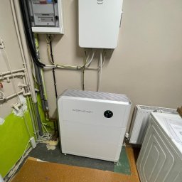 GREEN LEAF POWER SPÓŁKA Z OGRANICZONĄ ODPOWIEDZIALNOŚCIĄ - Biały magazyn energii SIDEnergy z podłączonymi kablami, zamontowany w pomieszczeniu z instalacją elektryczną i grzewczą. Widoczny licznik i zabezpieczenia.