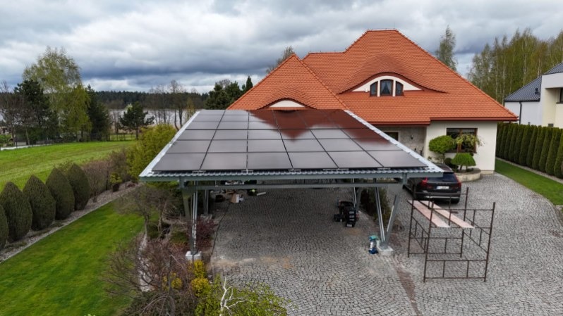 Zadaszenie na samochód z panelami słonecznymi na posesji z domem o czerwonym dachu i brukowanym podjazdem. Widoczne rusztowanie i auto.
