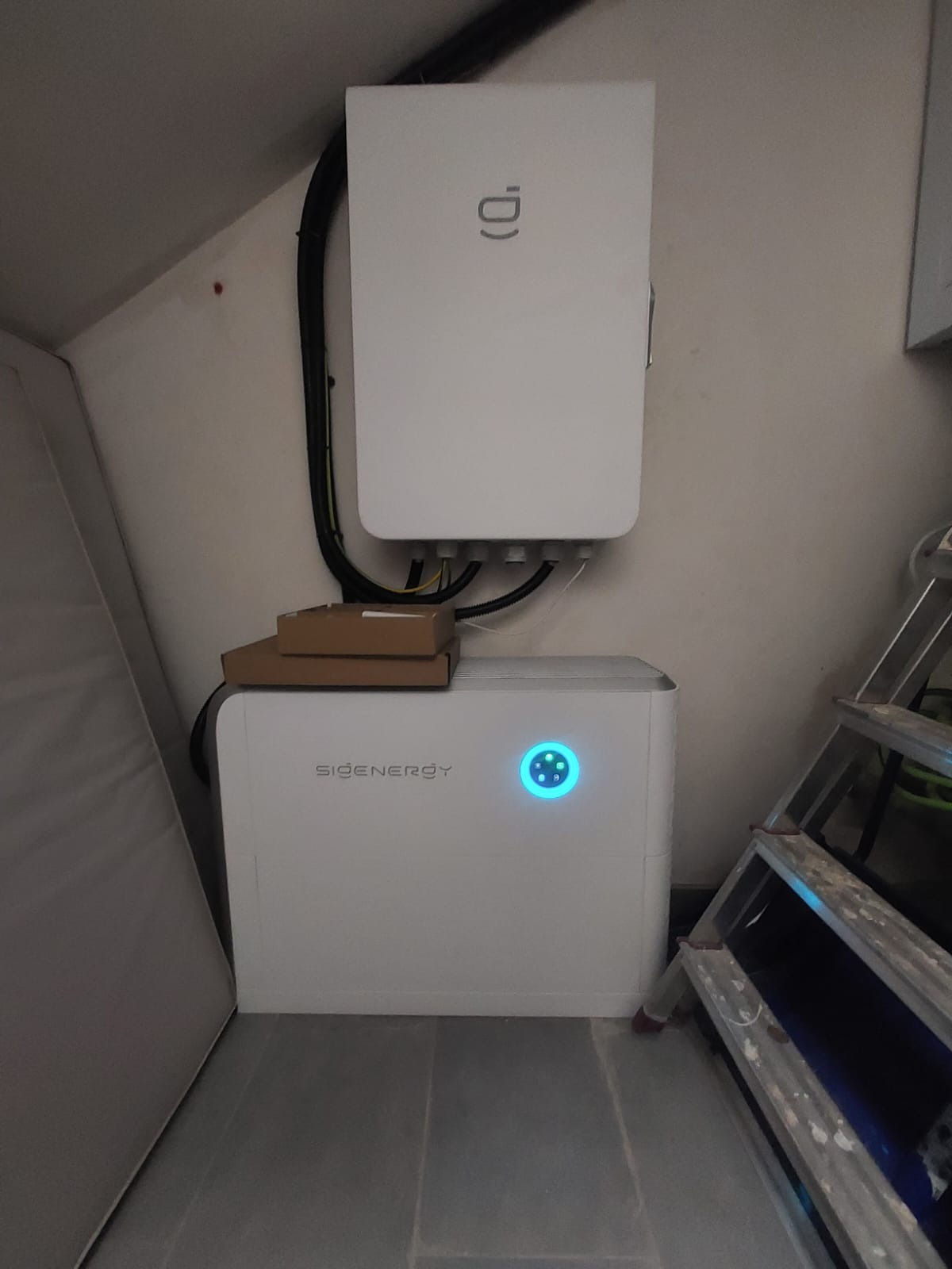 Biały akumulator energii SIGENERGY z niebieskim wskaźnikiem LED, zamontowany w pomieszczeniu, z widocznymi przewodami i drabiną.