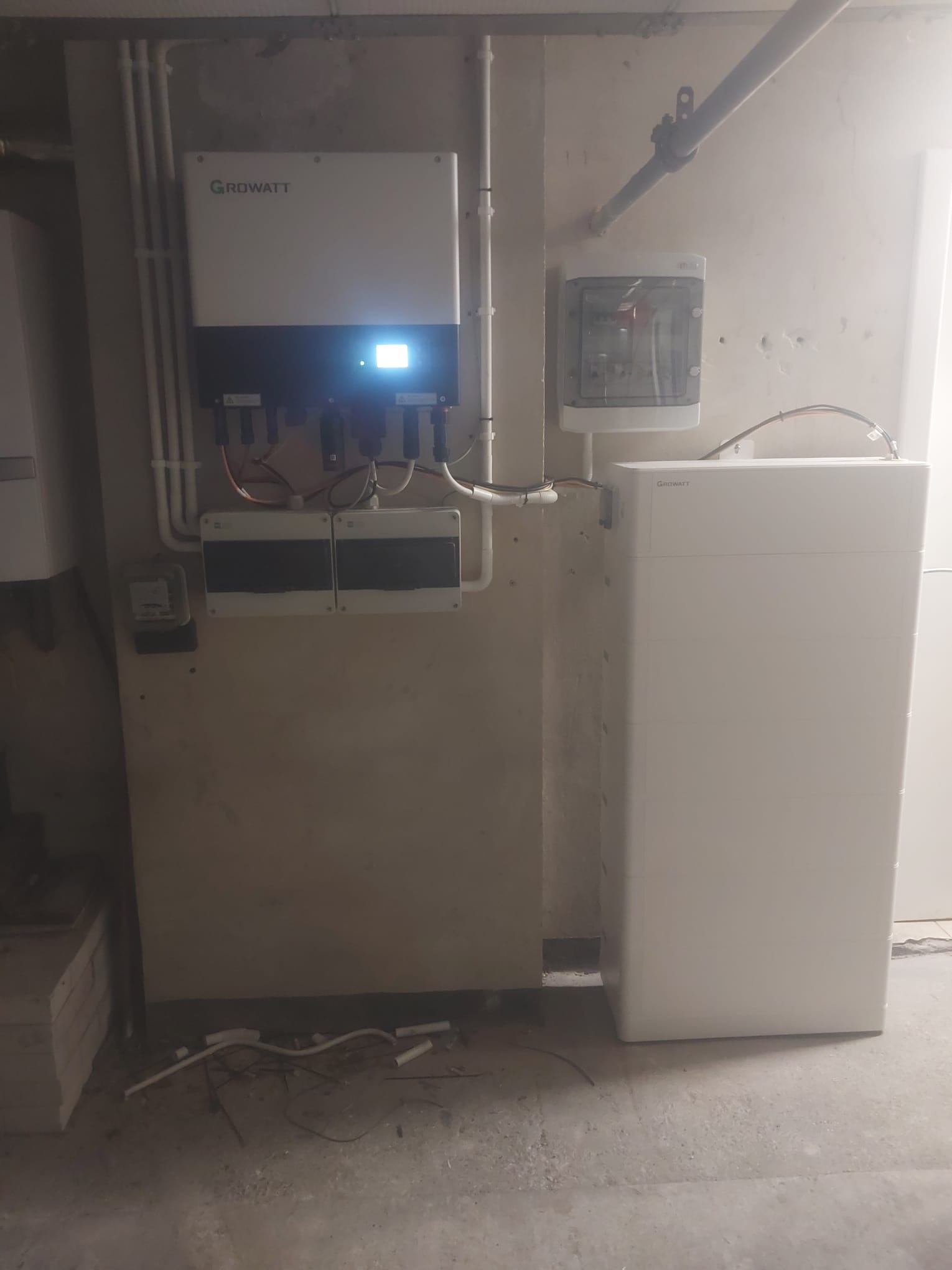 Zamontowany inwerter Growatt z podłączonymi przewodami, obok biały akumulator energii i skrzynka elektryczna na szarej ścianie w surowym pomieszczeniu.