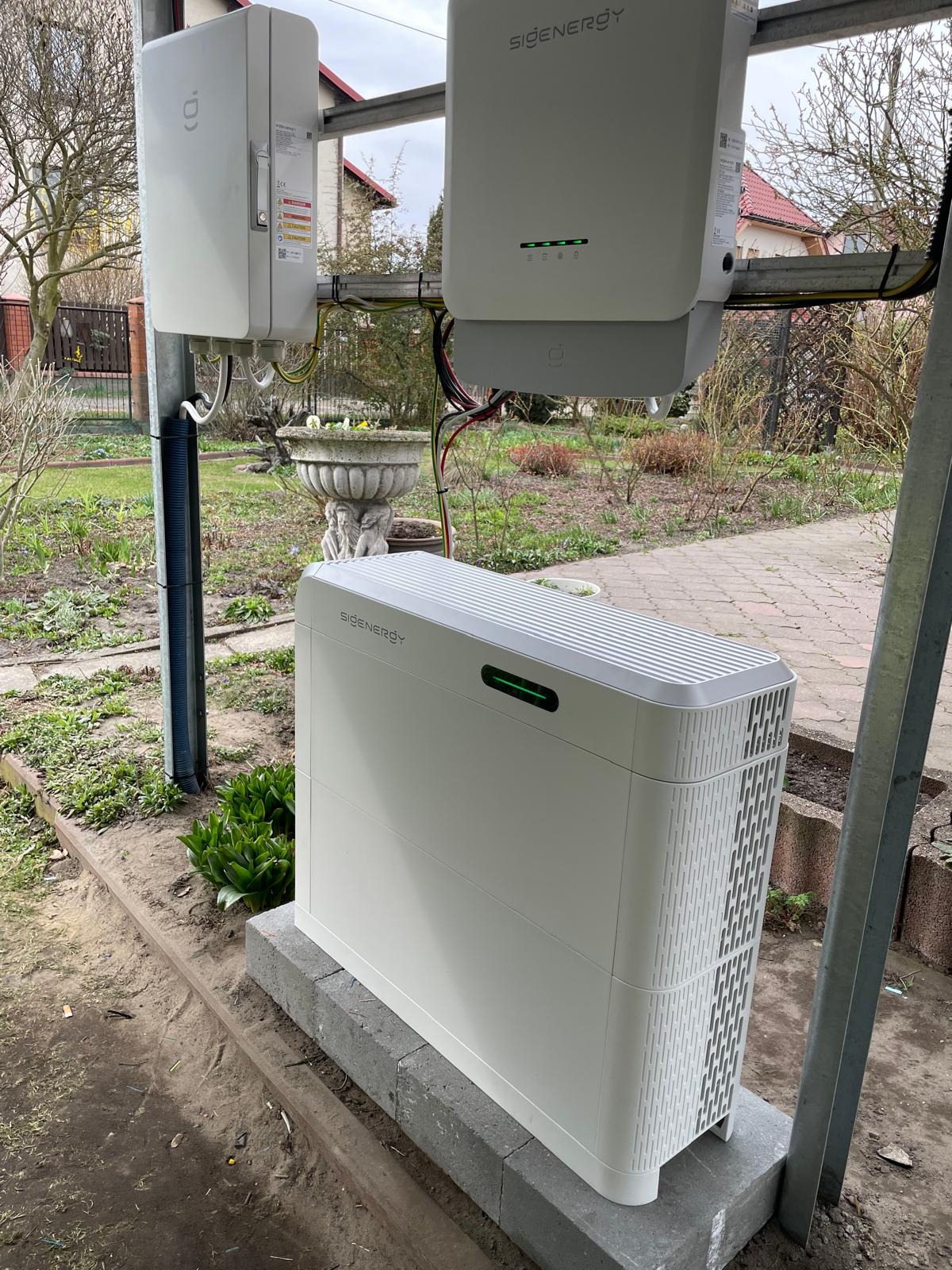 Biały, nowoczesny magazyn energii marki Sigenergy, zainstalowany na zewnątrz, z widocznymi elementami montażowymi i otoczeniem ogrodowym.