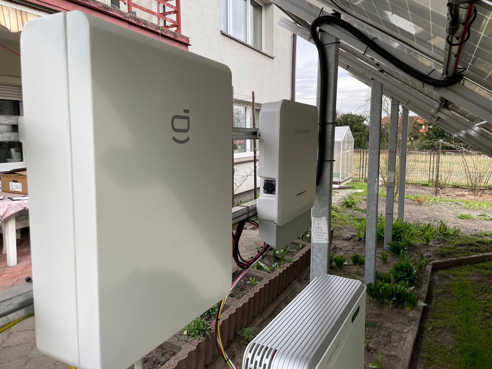 Białe obudowy urządzeń fotowoltaicznych zamontowane na zewnątrz budynku, widoczne panele słoneczne i elementy instalacji w ogrodzie.