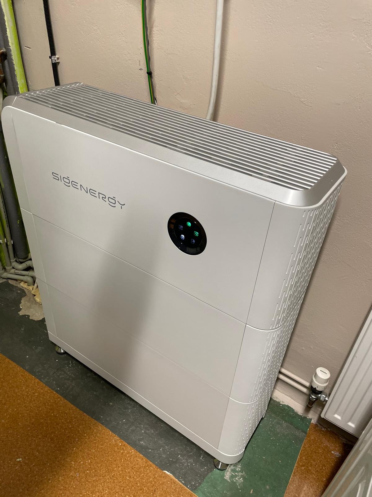 Biały, nowoczesny magazyn energii SiGenergy z okrągłym panelem sterowania, ustawiony w pomieszczeniu z widocznymi rurami i instalacjami na ścianie.