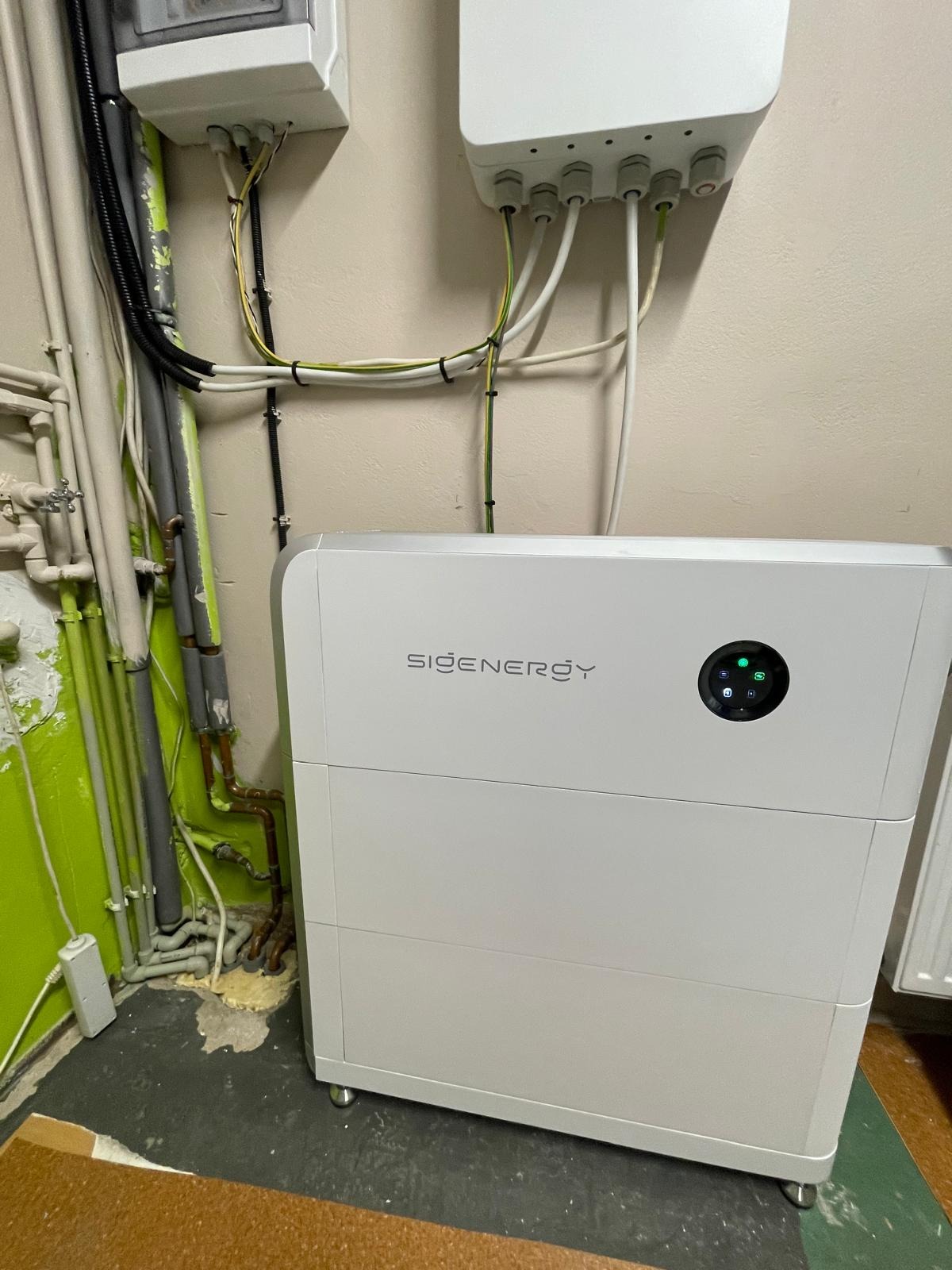 Biały magazyn energii Sienergy z podłączonymi kablami, umieszczony w pomieszczeniu z rurami i instalacjami elektrycznymi. Widoczne diody sygnalizacyjne.
