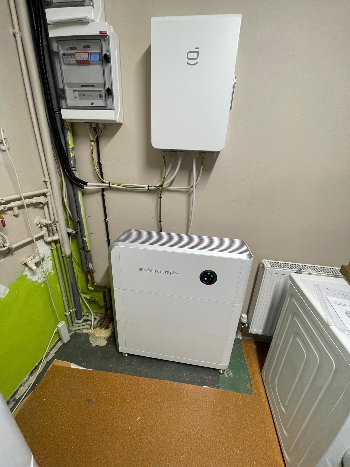 Biały magazyn energii SIDEnergy z podłączonymi kablami, zamontowany w pomieszczeniu z instalacją elektryczną i grzewczą. Widoczny licznik i zabezpieczenia.