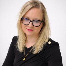 KANCELARIA ADWOKATA I DORADCY PODATKOWEGO ALEKSANDRA MEDLARSKA - Portret kobiety z blond włosami, w czarnej sukience z ozdobnymi guzikami i okularach, na jasnym tle. Uśmiech, delikatny makijaż, złoty naszyjnik.