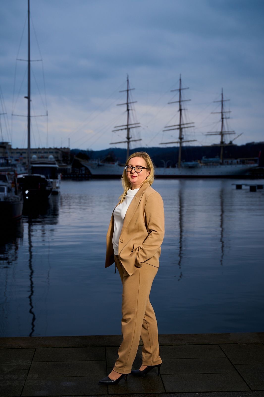Kobieta w beżowym garniturze i okularach, stojąca na tle portu z żaglowcami. Profesjonalny wizerunek na tle wody i statków.