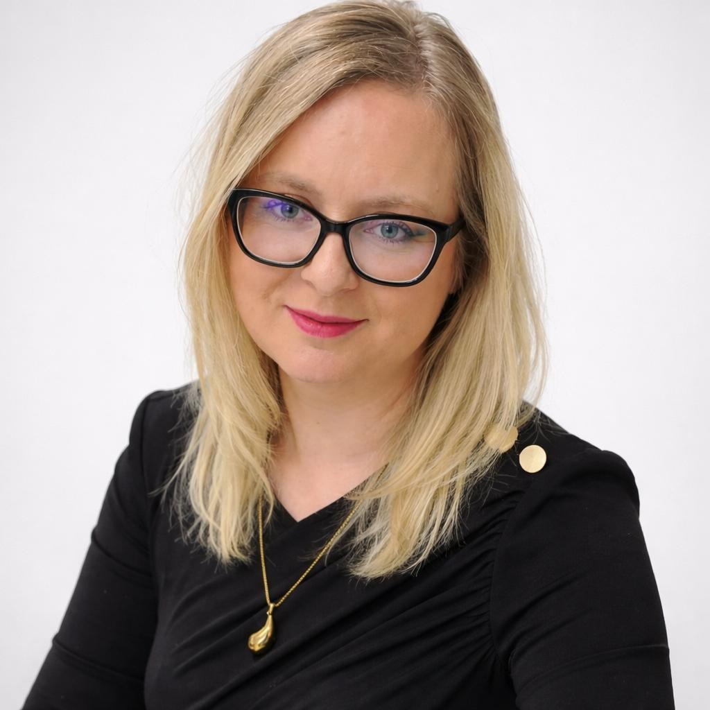 Portret kobiety z blond włosami, w czarnej sukience z ozdobnymi guzikami i okularach, na jasnym tle. Uśmiech, delikatny makijaż, złoty naszyjnik.