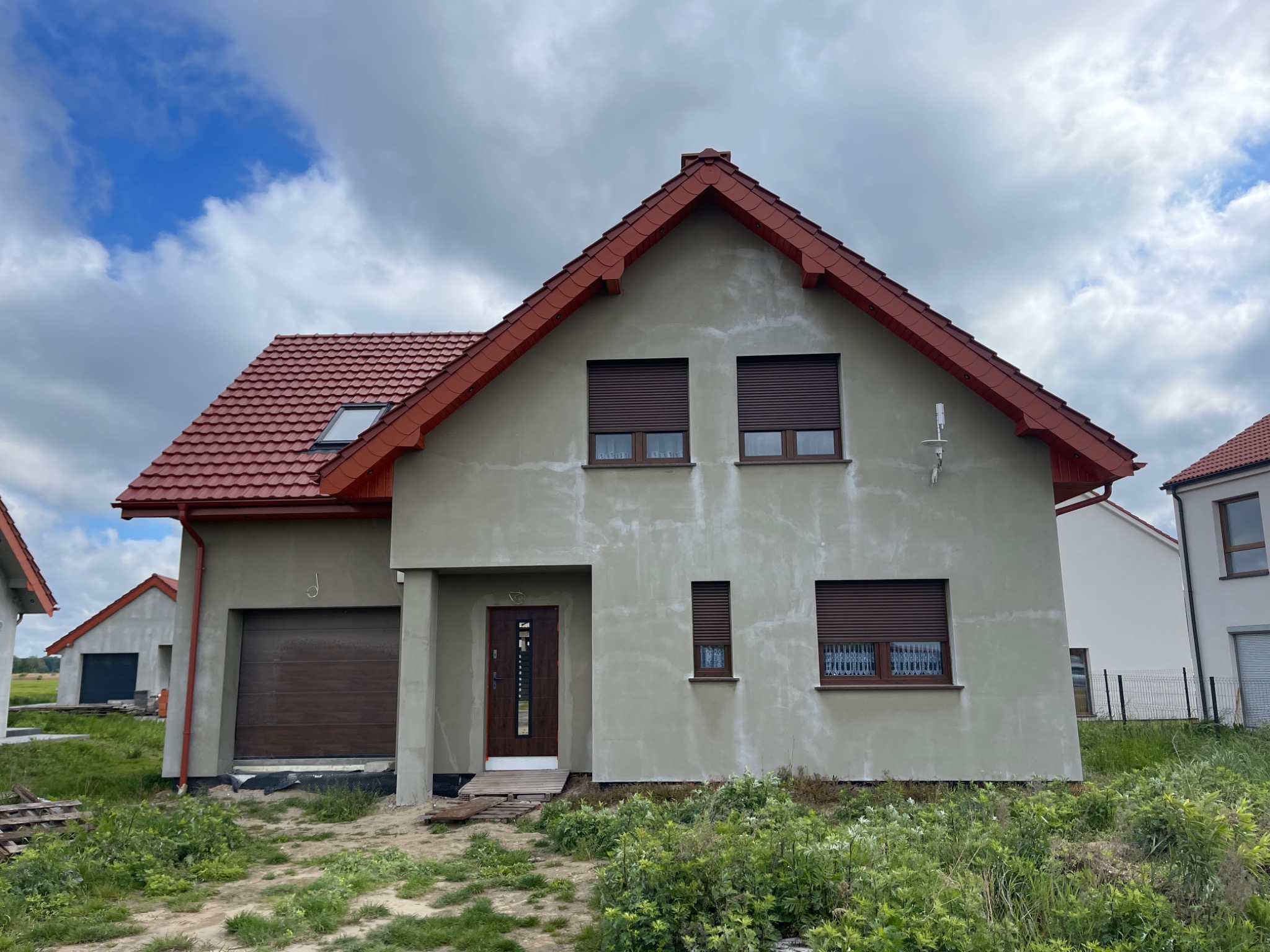 Dom jednorodzinny z czerwoną dachówką i brązowymi roletami, ściany w trakcie wykończenia z widocznymi łatami. Widok z poziomu gruntu, pochmurne niebo.