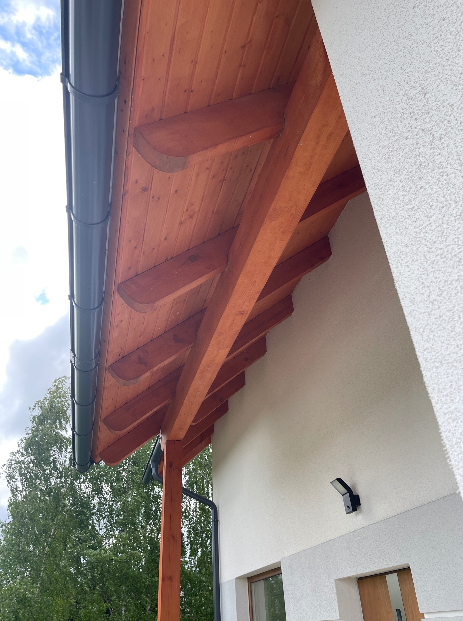 Drewniana podbitka dachowa z widocznymi belkami i zamontowaną rynną w kolorze grafitowym, na tle białej elewacji budynku. Widok z dołu.