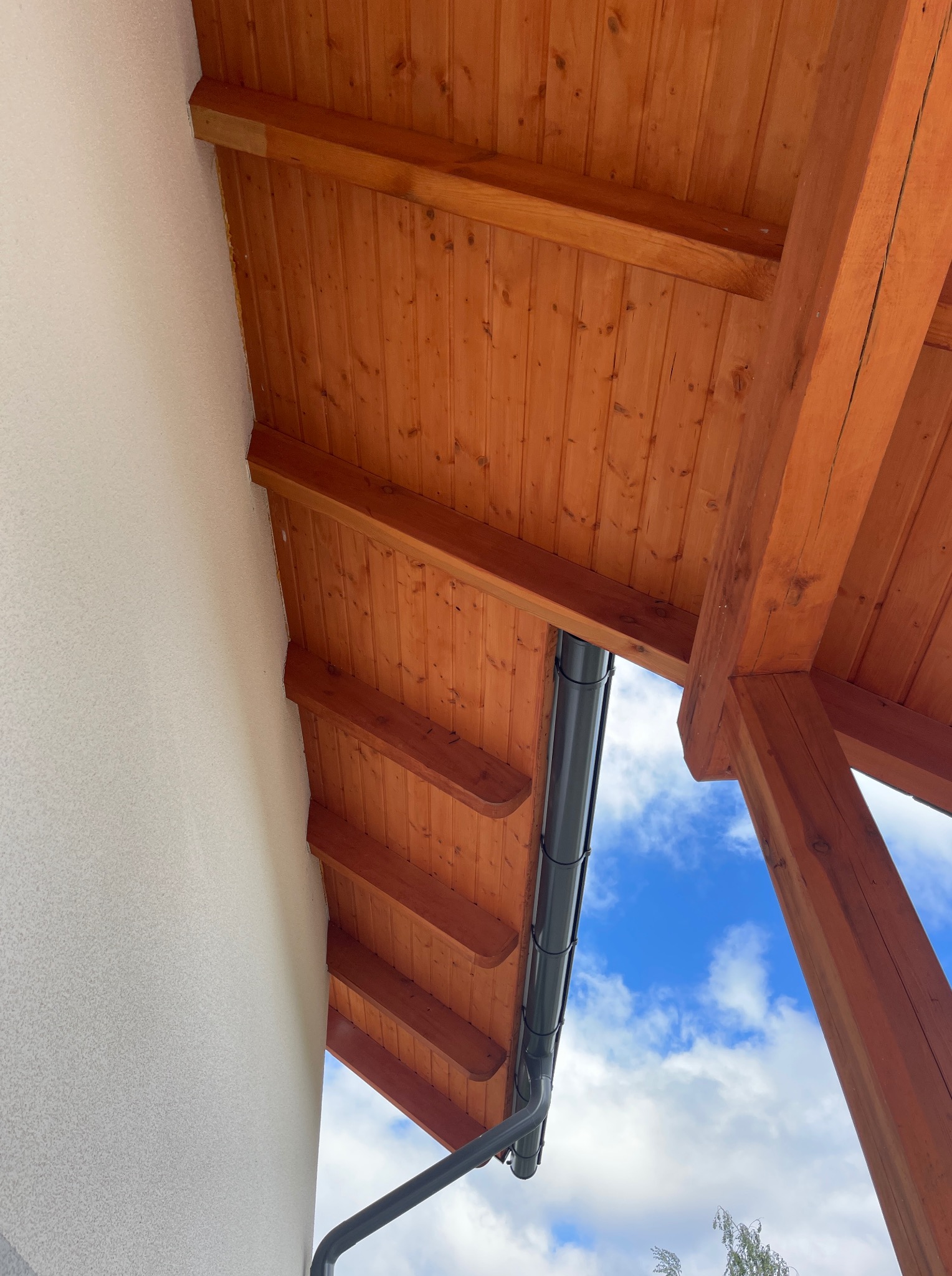 Drewniana podbitka dachowa z widoczną rynną w kolorze grafitowym na tle błękitnego nieba. Ujęcie z dołu, podkreślające detale konstrukcji.