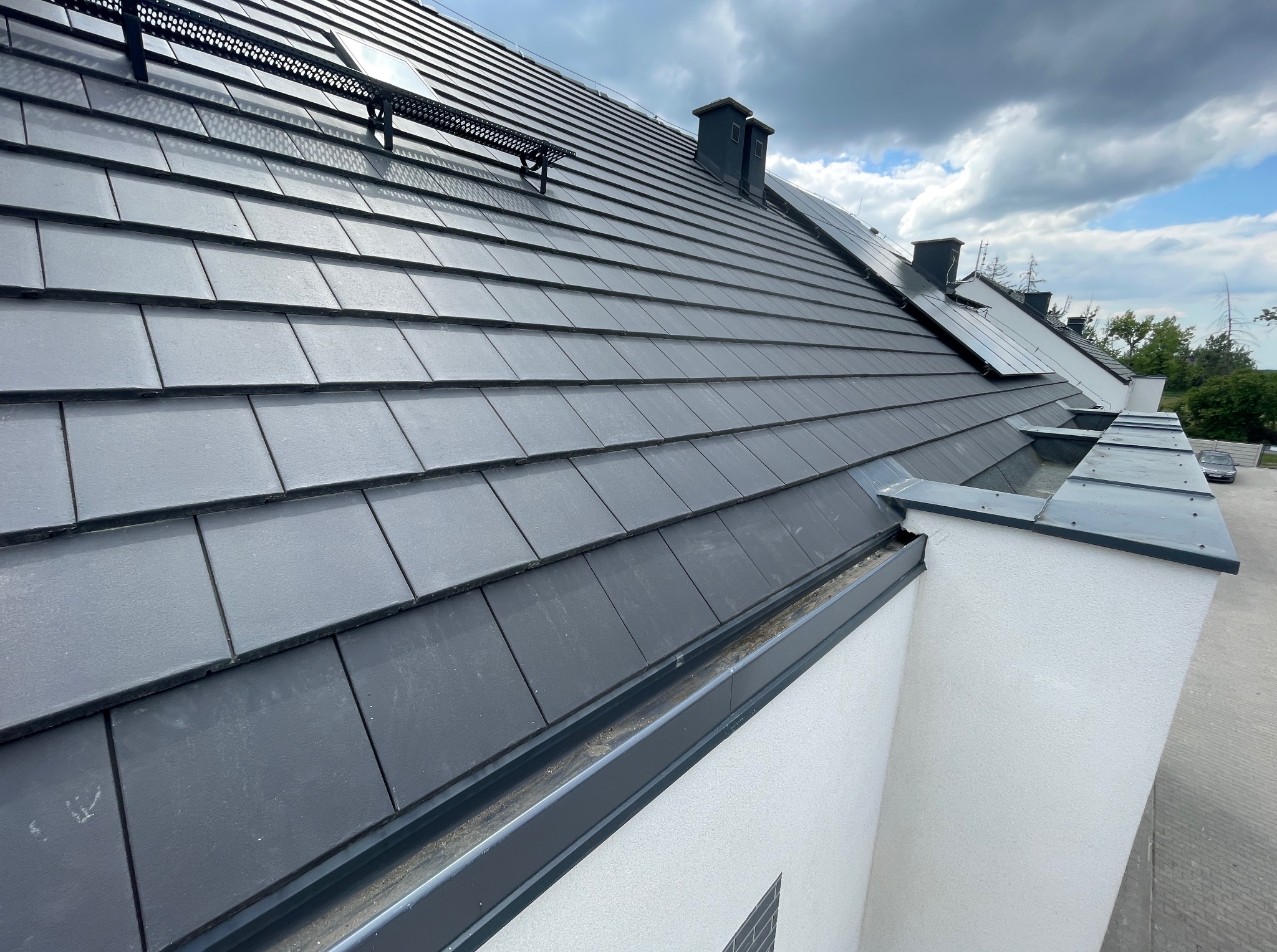 Szary dach z dachówki ceramicznej, widok z góry, z zamontowanymi zabezpieczeniami śniegowymi i obróbkami blacharskimi w kolorze grafitowym. Widoczne kominy i fragment elewacji.