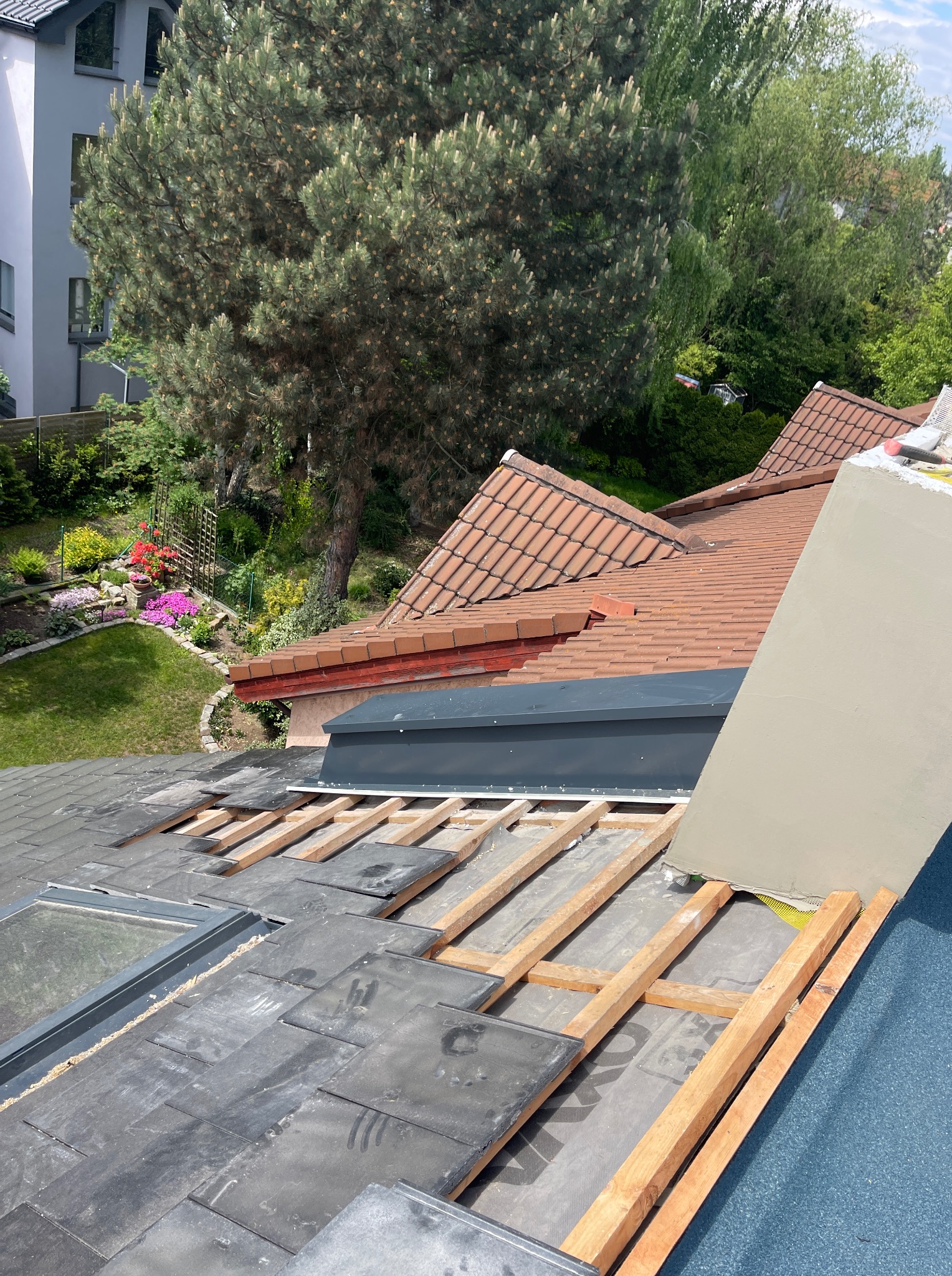 Fragment dachu w trakcie wymiany z widocznym oknem dachowym i drewnianą konstrukcją. W tle ogród z kwiatami i drzewami.