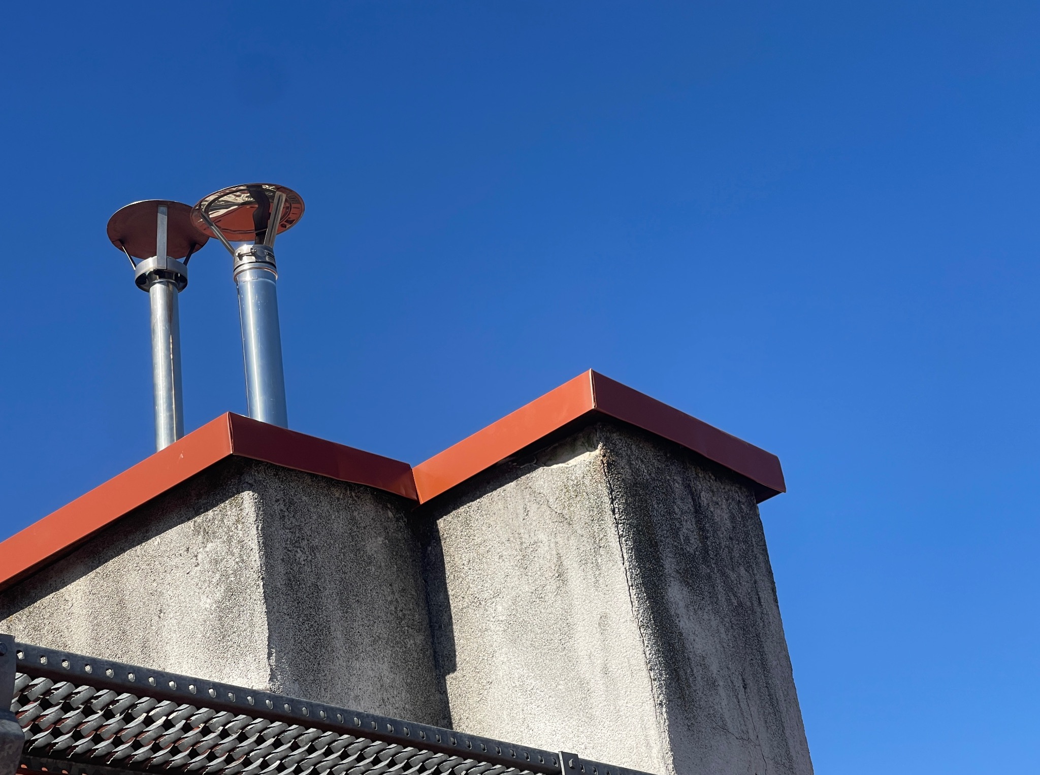 Dwa kominy wentylacyjne z metalowymi rurami i czerwonymi daszkami, widziane z dołu na tle błękitnego nieba. Fragment metalowej konstrukcji na pierwszym planie.