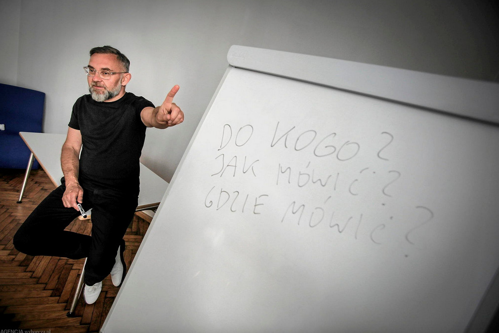 Mężczyzna w czarnym ubraniu prezentuje pytania na flipcharcie: 'Do kogo? Jak mówić? Gdzie mówić?' podczas szkolenia. Dłoń z markerem, białe buty, jasne tło.