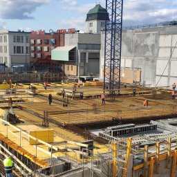 Mykhailo Olenich - Prace zbrojarskie na budowie w Krakowie: widok z góry na przygotowane zbrojenie fundamentów, pracownicy w pomarańczowych kamizelkach, szalunki i dźwig w tle.