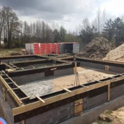 Częściowo wylane fundamenty budynku z widocznym zbrojeniem, szalunkiem, izolacją i bloczkami betonowymi. Na drugim planie hałdy piachu i materiały budowlane.