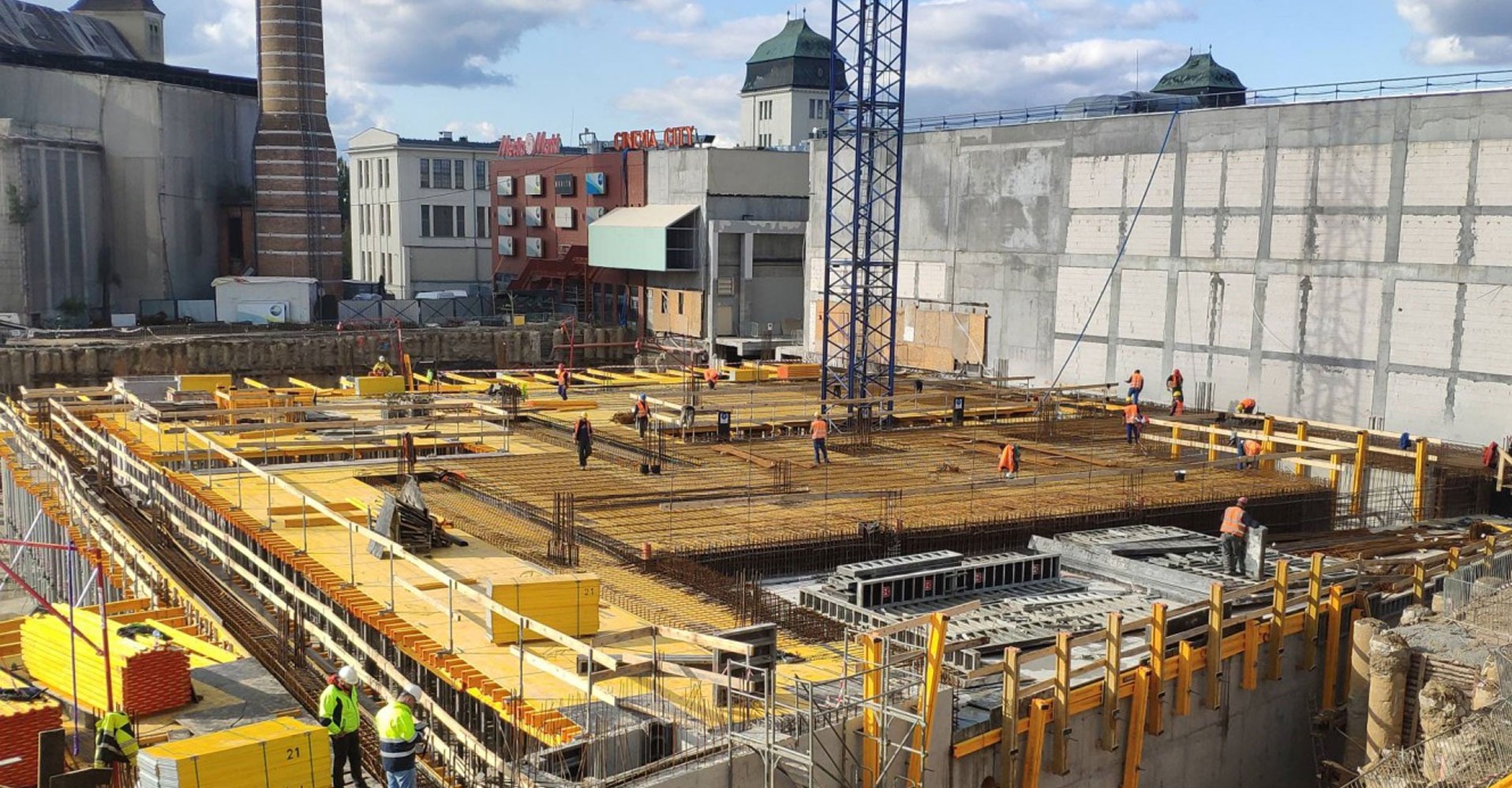 Prace zbrojarskie na budowie w Krakowie: widok z góry na przygotowane zbrojenie fundamentów, pracownicy w pomarańczowych kamizelkach, szalunki i dźwig w tle.