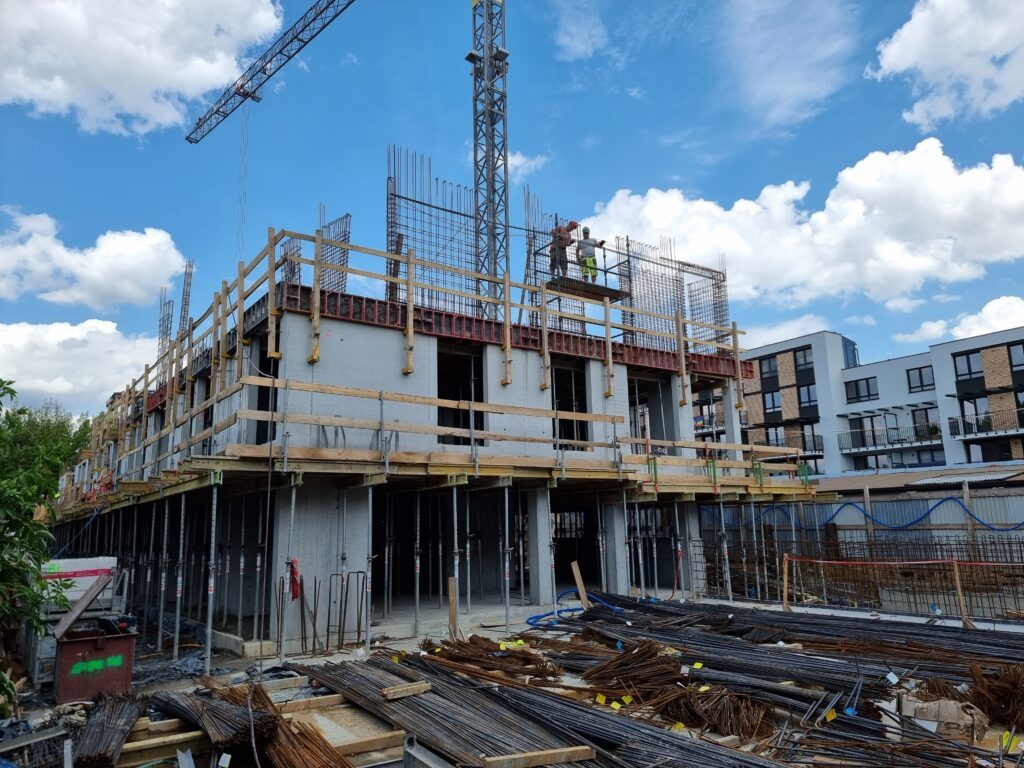 Wznoszenie budynku żelbetowego: szalunki, zbrojenia, pracownicy na rusztowaniu. W tle błękitne niebo z chmurami i fragmenty zabudowy wielorodzinnej.