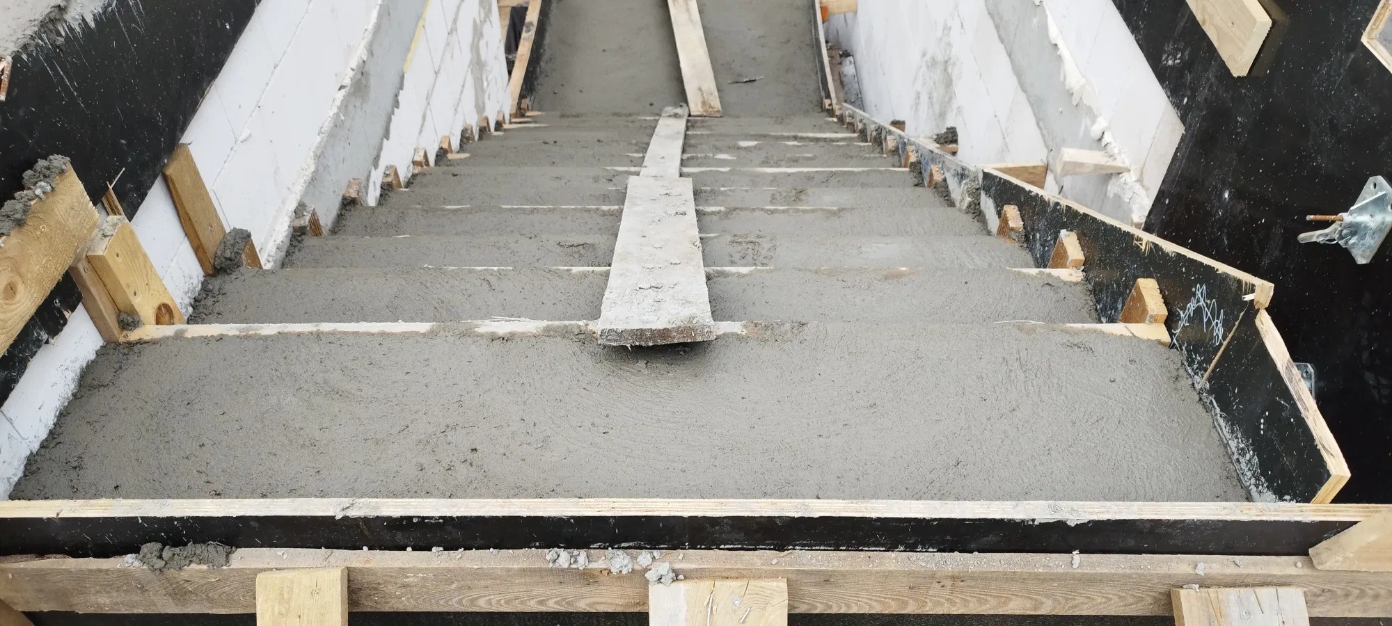 Surowe schody betonowe w szalunku, widok z góry. Świeża wylewka pokrywa stopnie, drewniane deski stabilizują konstrukcję. Etap budowy, surowy beton.