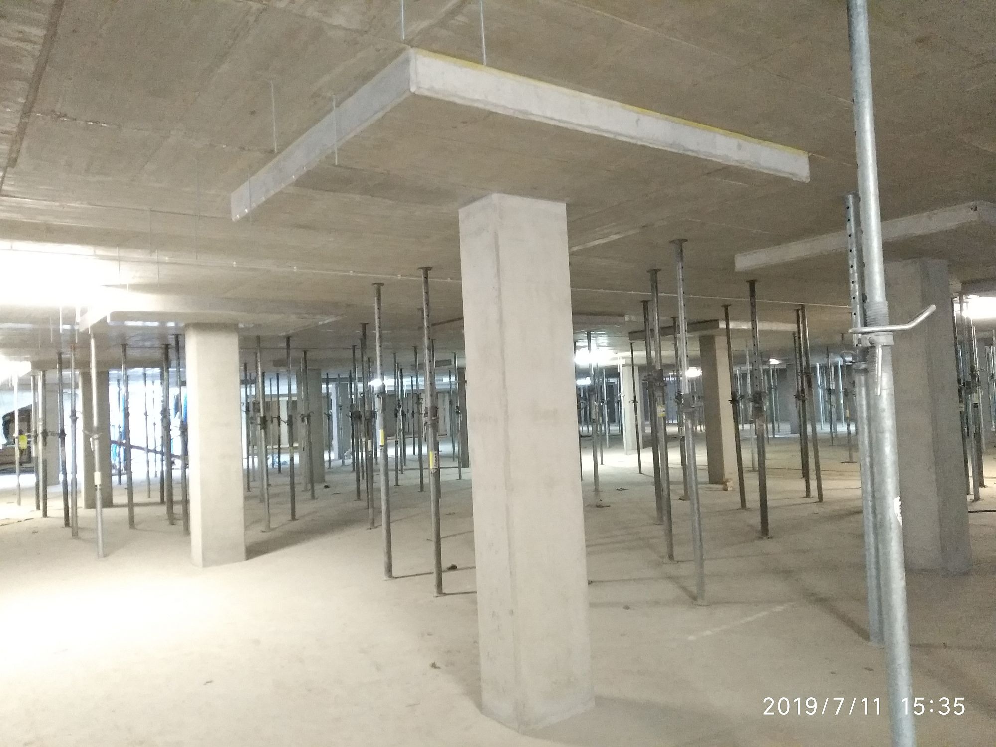 Betonowe filary i podpory stropu na placu budowy. Widoczne szare elementy konstrukcyjne i metalowe stemple podtrzymujące strop. Surowy stan, brak wykończenia.
