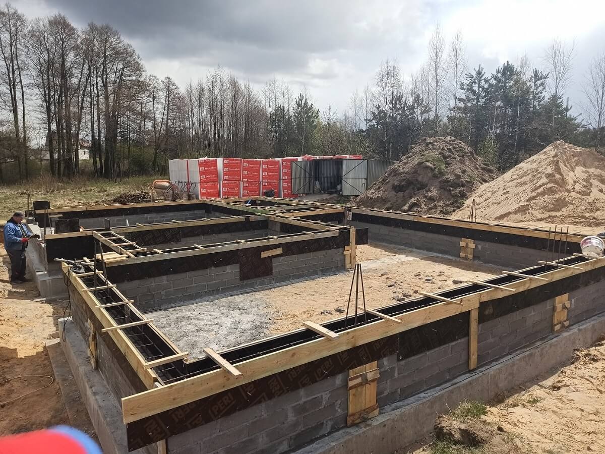 Częściowo wylane fundamenty budynku z widocznym zbrojeniem, szalunkiem, izolacją i bloczkami betonowymi. Na drugim planie hałdy piachu i materiały budowlane.