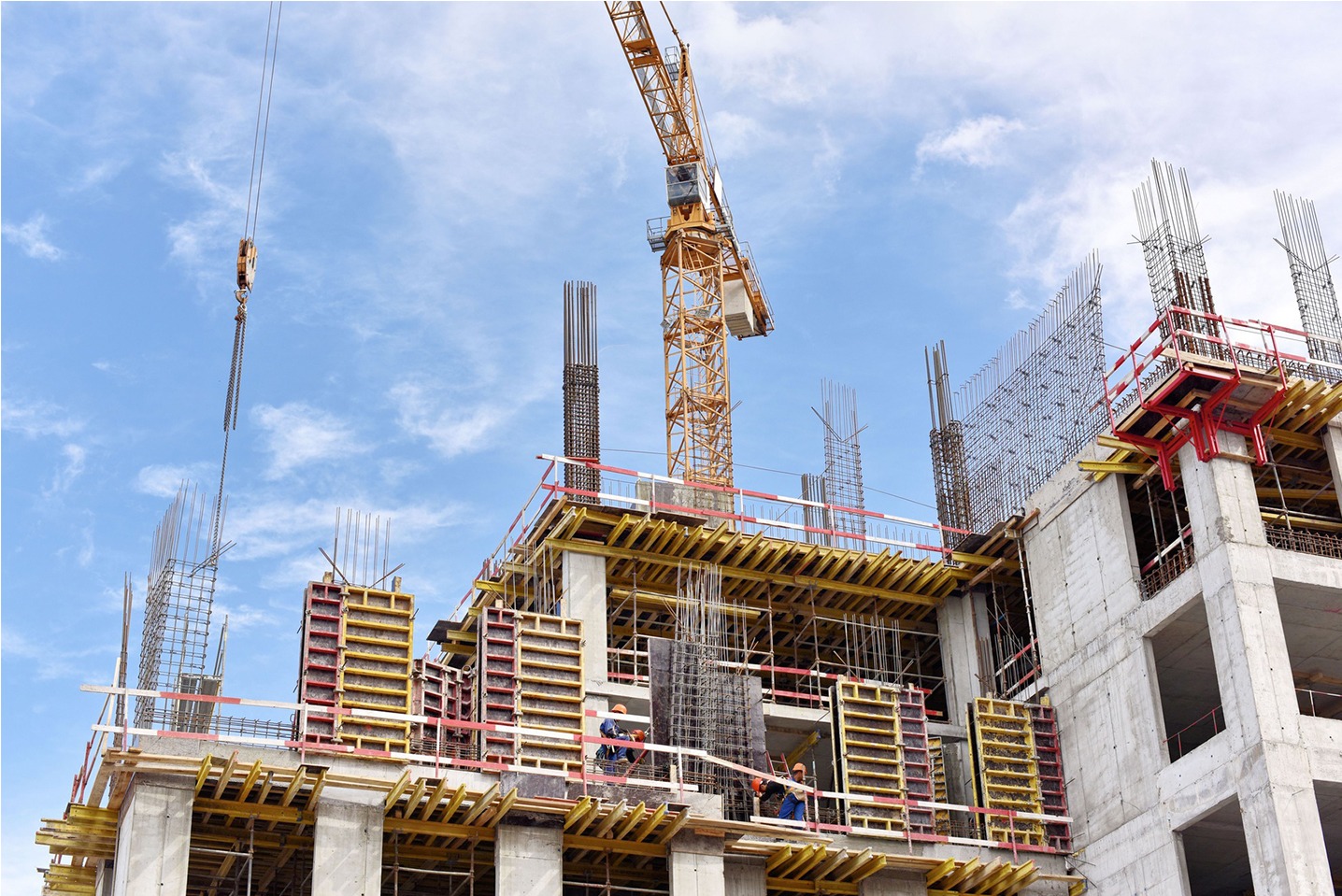 Wysoki budynek w trakcie budowy, widoczne zbrojenia, szalunki, dźwig i pracujący robotnicy w kaskach na tle błękitnego nieba z chmurami.