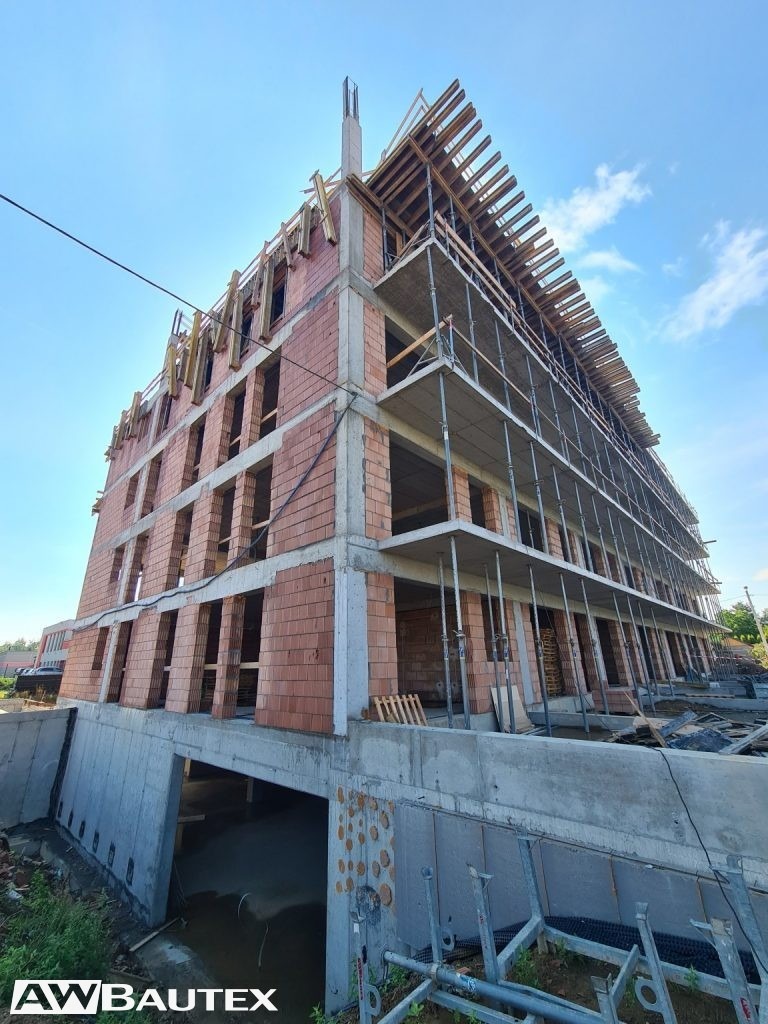 Wysoki budynek w trakcie budowy, z widoczną konstrukcją szkieletową, ceglanymi ścianami i betonowym fundamentem. Ujęcie z dołu, błękitne niebo w tle.