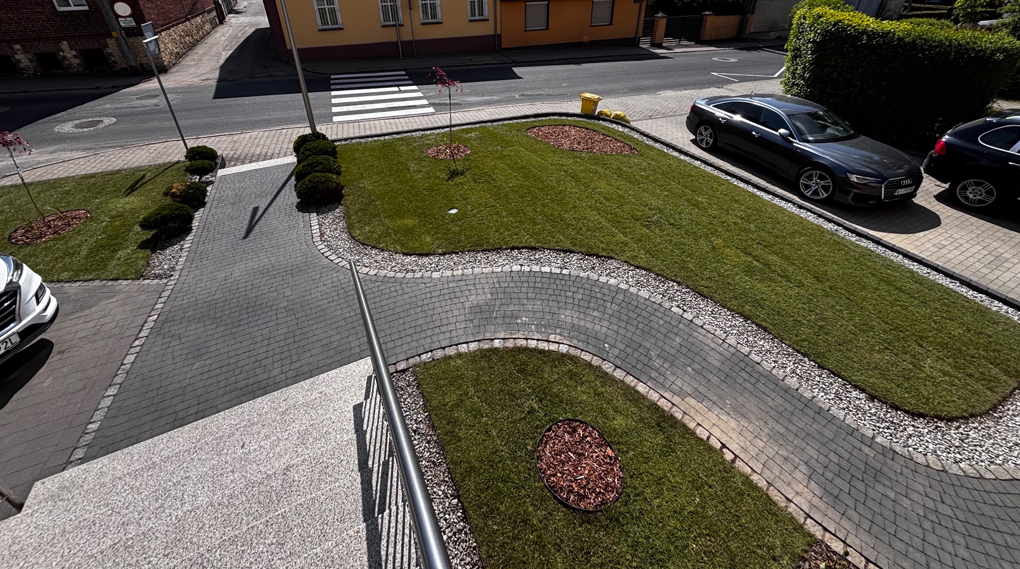 Widok z góry na świeżo założony trawnik z rolki, obrzeża z kamienia i kostki, z elementami dekoracyjnymi z kory. W tle zaparkowane samochody i zabudowa miejska.