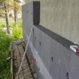 Ocieplanie elewacji: fragment ściany z szarym styropianem, widoczne otwory i pomarańczowe znaczniki. Częściowo ocieplona ściana budynku. Na dole kostka brukowa i roślinność.