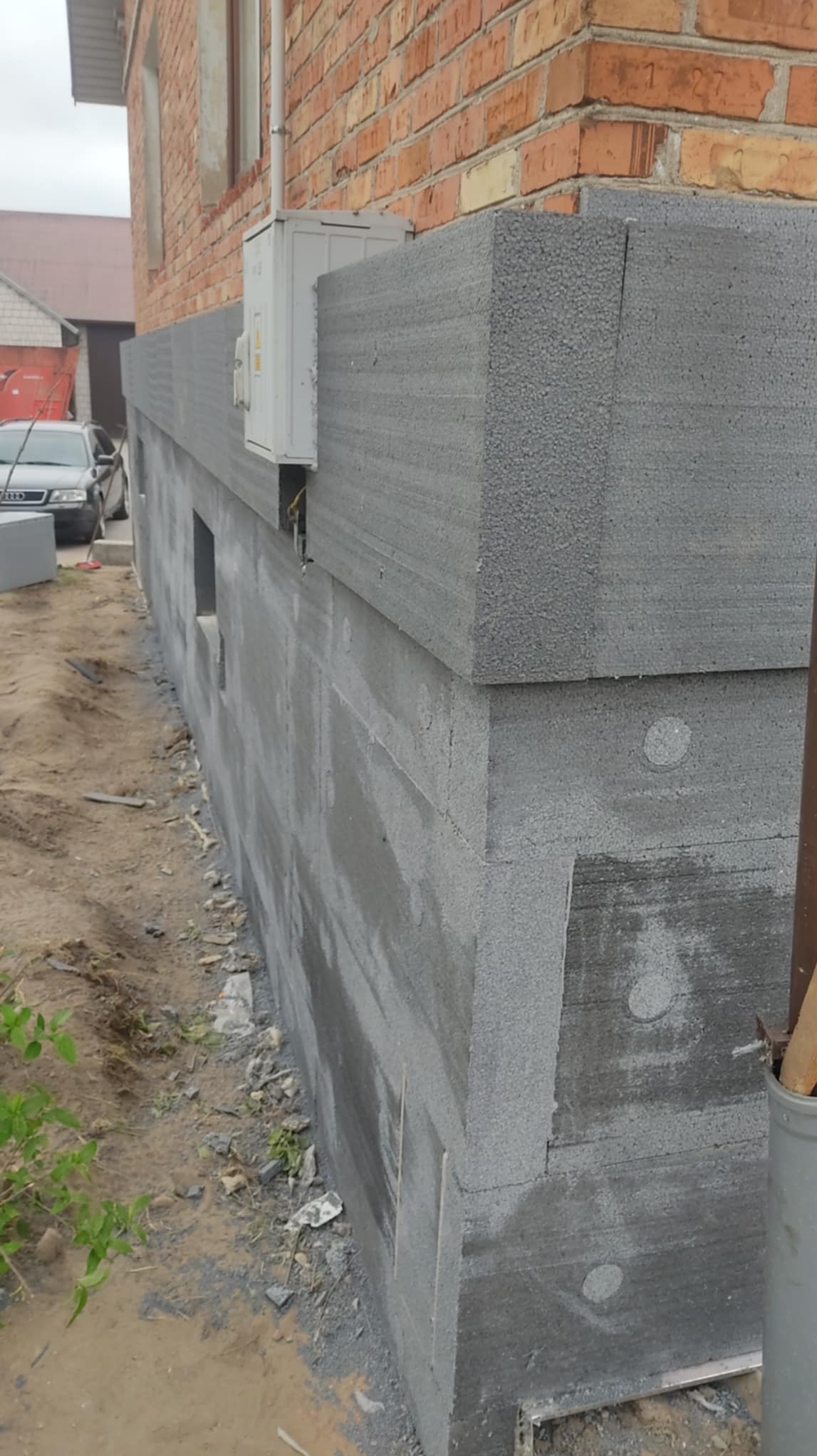 Ocieplony fundament budynku z bloków styropianowych, widoczna ściana z czerwonej cegły i klimatyzator. Etap prac ociepleniowych, szara kolorystyka.
