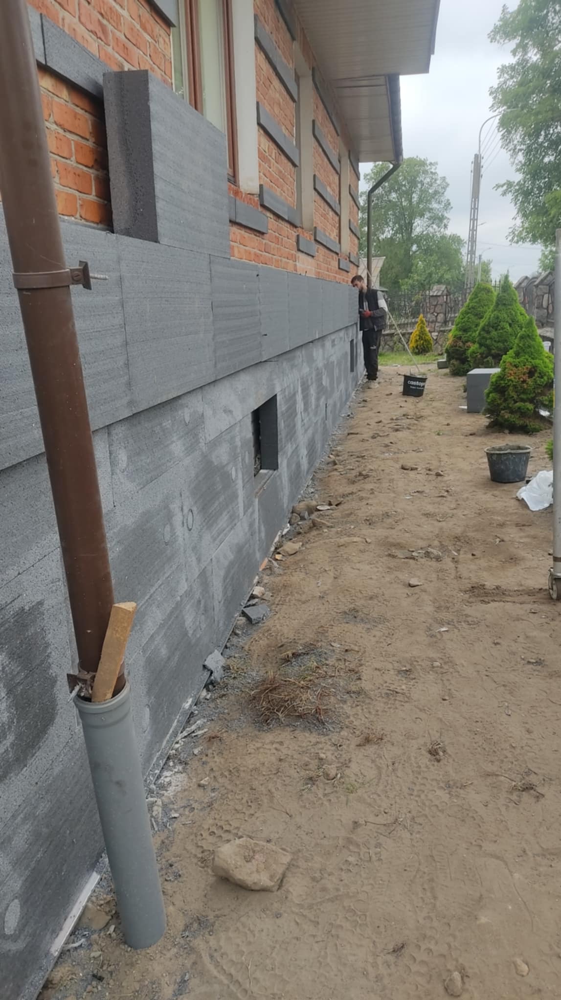 Ocieplanie elewacji styropianem grafitowym w Sokołowie Podlaskim. Widoczny fragment ściany z oknem. Pracownik w tle. Etap przed nałożeniem tynku.