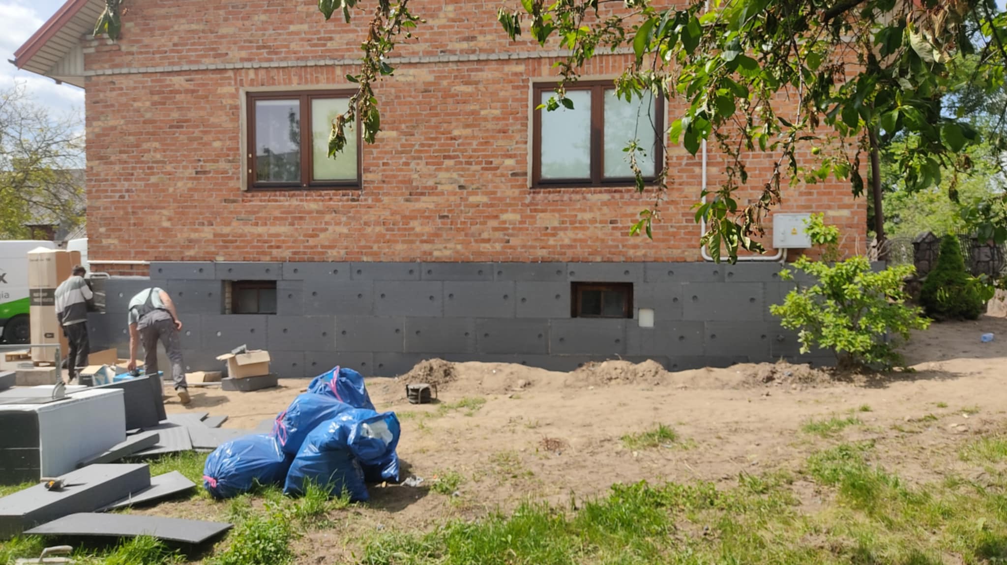 Ocieplanie fundamentów domu z cegły. Pracownicy montują szare płyty izolacyjne, widoczne okna piwniczne i worki z odpadami budowlanymi. Prace w toku.
