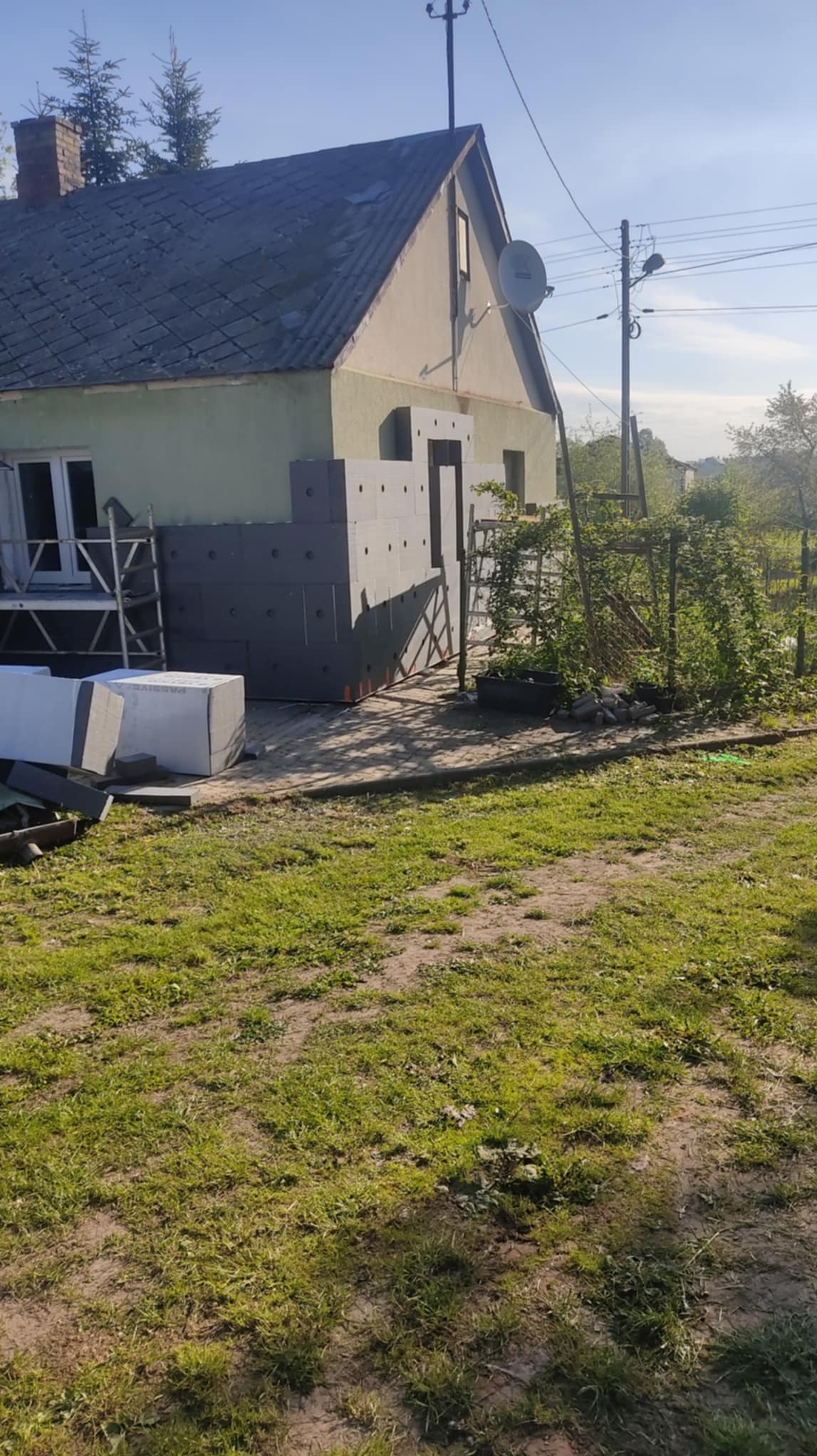 Ocieplanie domu szarym styropianem grafitowym. Widoczne płyty styropianowe i rusztowania. Dom z szarą dachówką i zieloną elewacją w trakcie prac ociepleniowych.