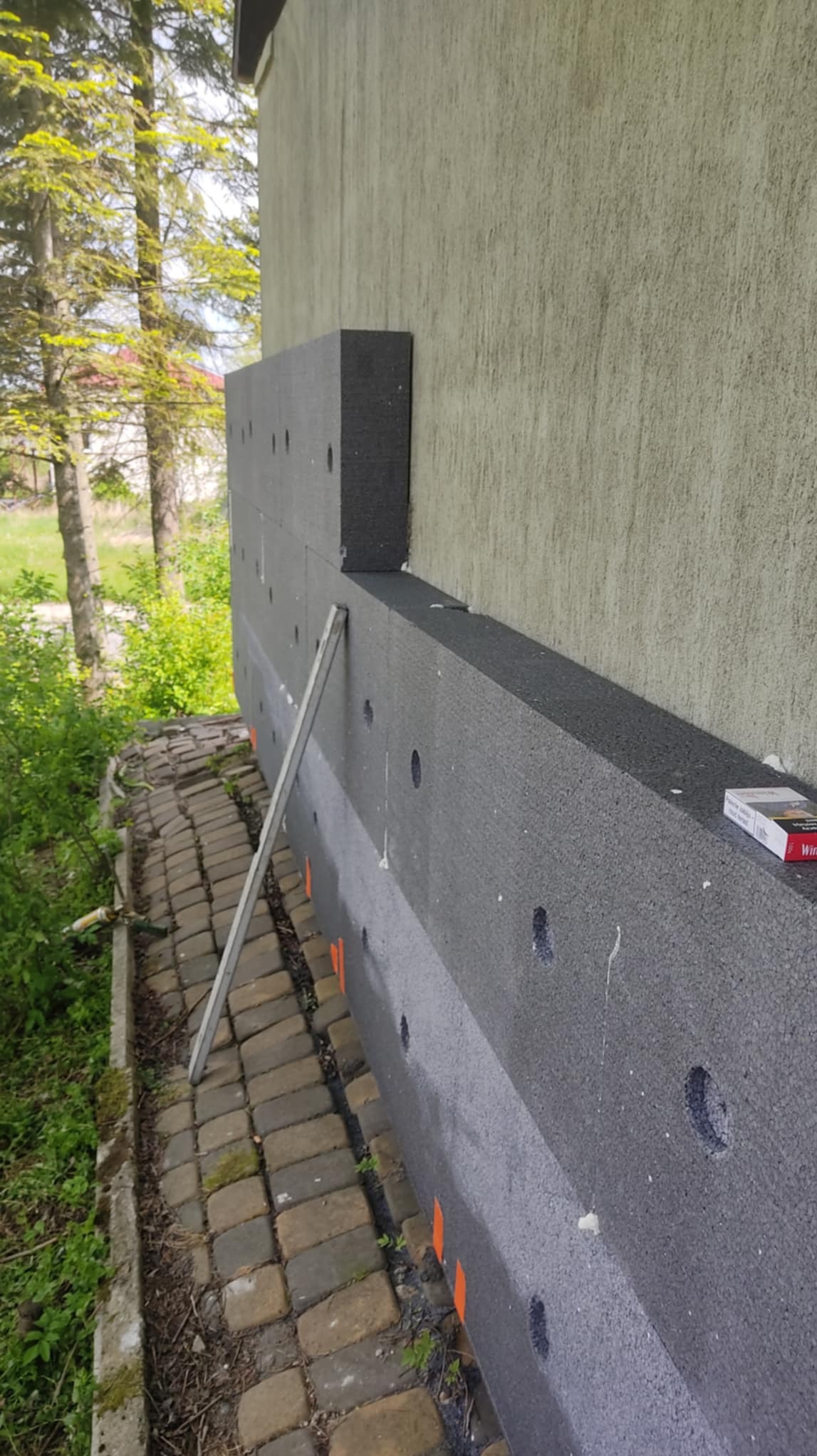 Ocieplanie elewacji: fragment ściany z szarym styropianem, widoczne otwory i pomarańczowe znaczniki. Częściowo ocieplona ściana budynku. Na dole kostka brukowa i roślinność.