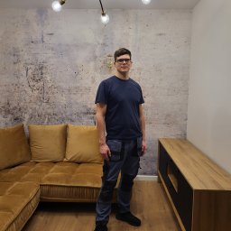 Adrian Zaliwski - Mężczyzna w odnowionym wnętrzu z ścianą o fakturze betonu, złotym narożnikiem i lampą w stylu retro, stojący na parkiecie.