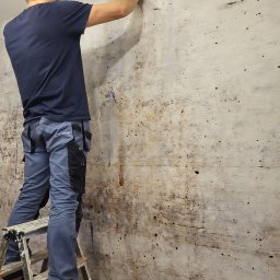 Adrian Zaliwski - Pracownik na drabinie szpachluje ścianę w pomieszczeniu. Widoczne ślady po poprzednich pracach. Ubrany w spodnie robocze z kieszeniami i ciemną koszulkę.